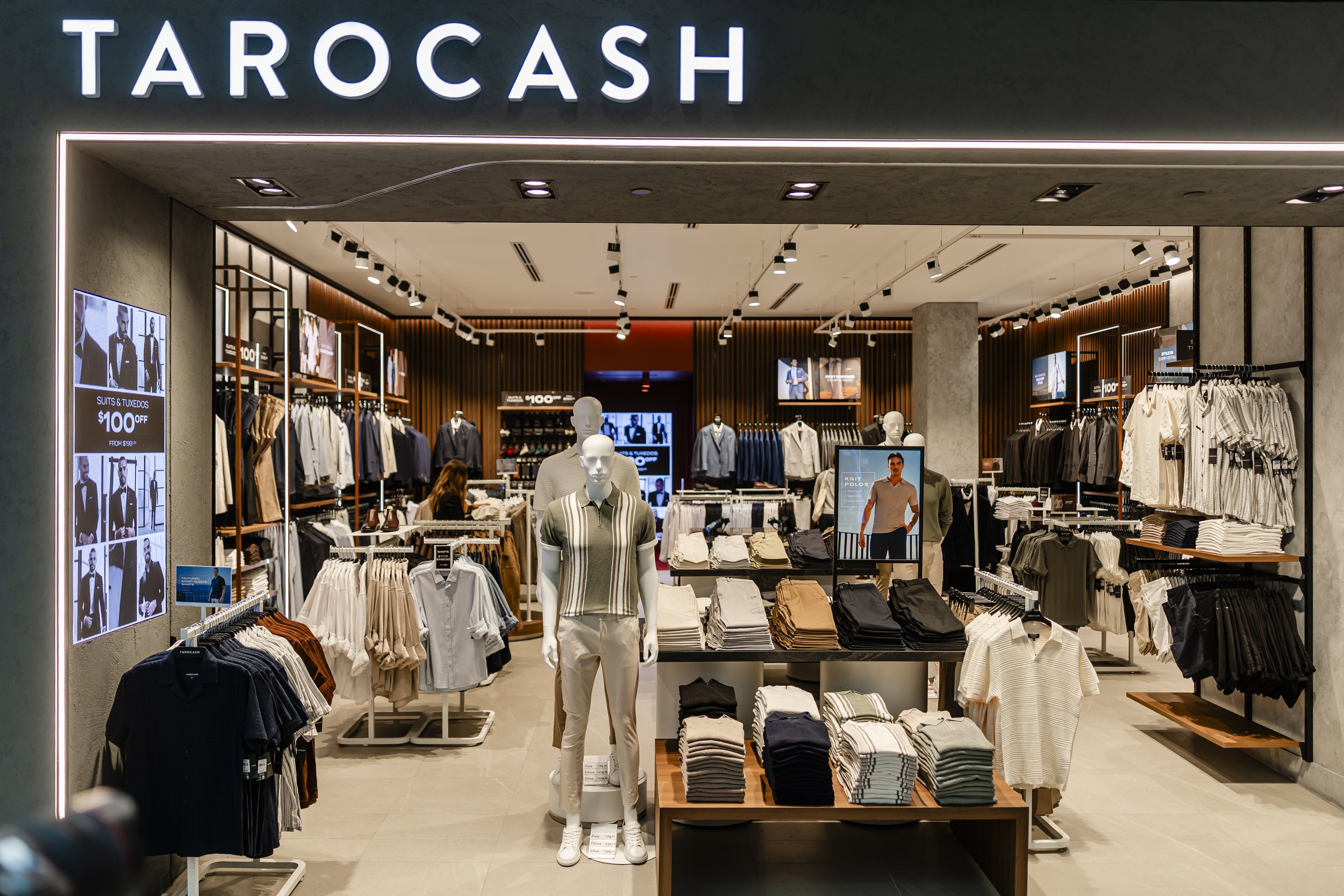 Clothing store interior with racks of men's and women's apparel, mannequins dressed in casual and business attire, and digital promotional displays showing discounts.