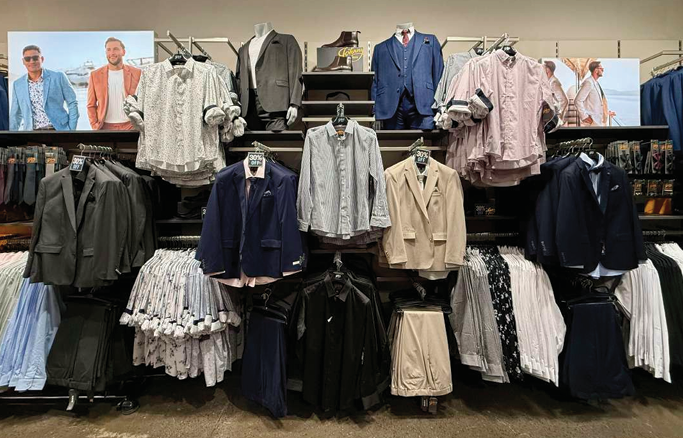 Clothing display in a retail store featuring men's and women's blazers, shirts, and skirts arranged on racks and shelves.