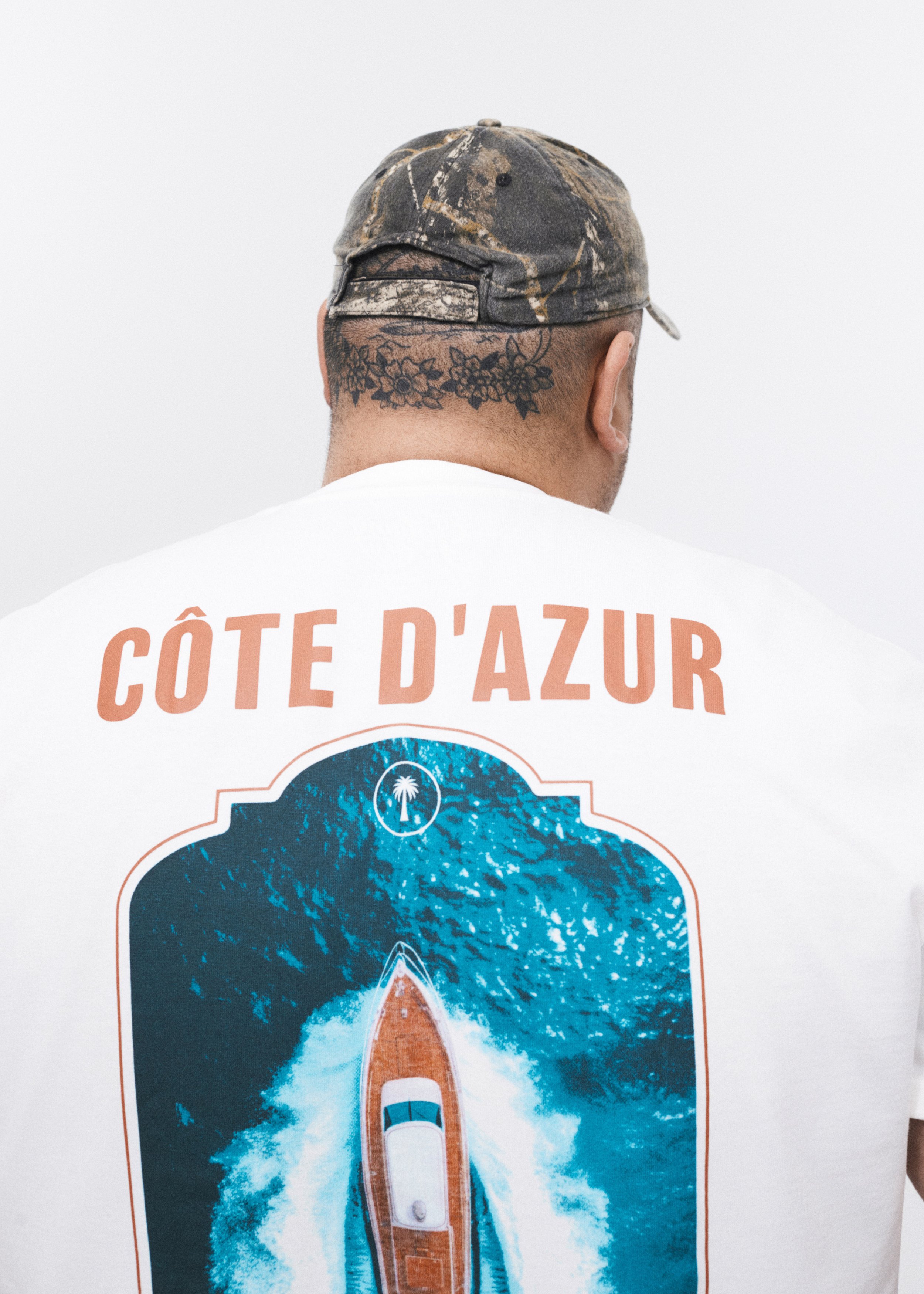 The back of a person wearing a camouflage cap and a white shirt with orange text, standing against a white background. The shirt reads "CÔTE D’AZUR." The person's head has a floral tattoo on the neck, and their back features a graphic design of a blue ocean wave with a road and a car.