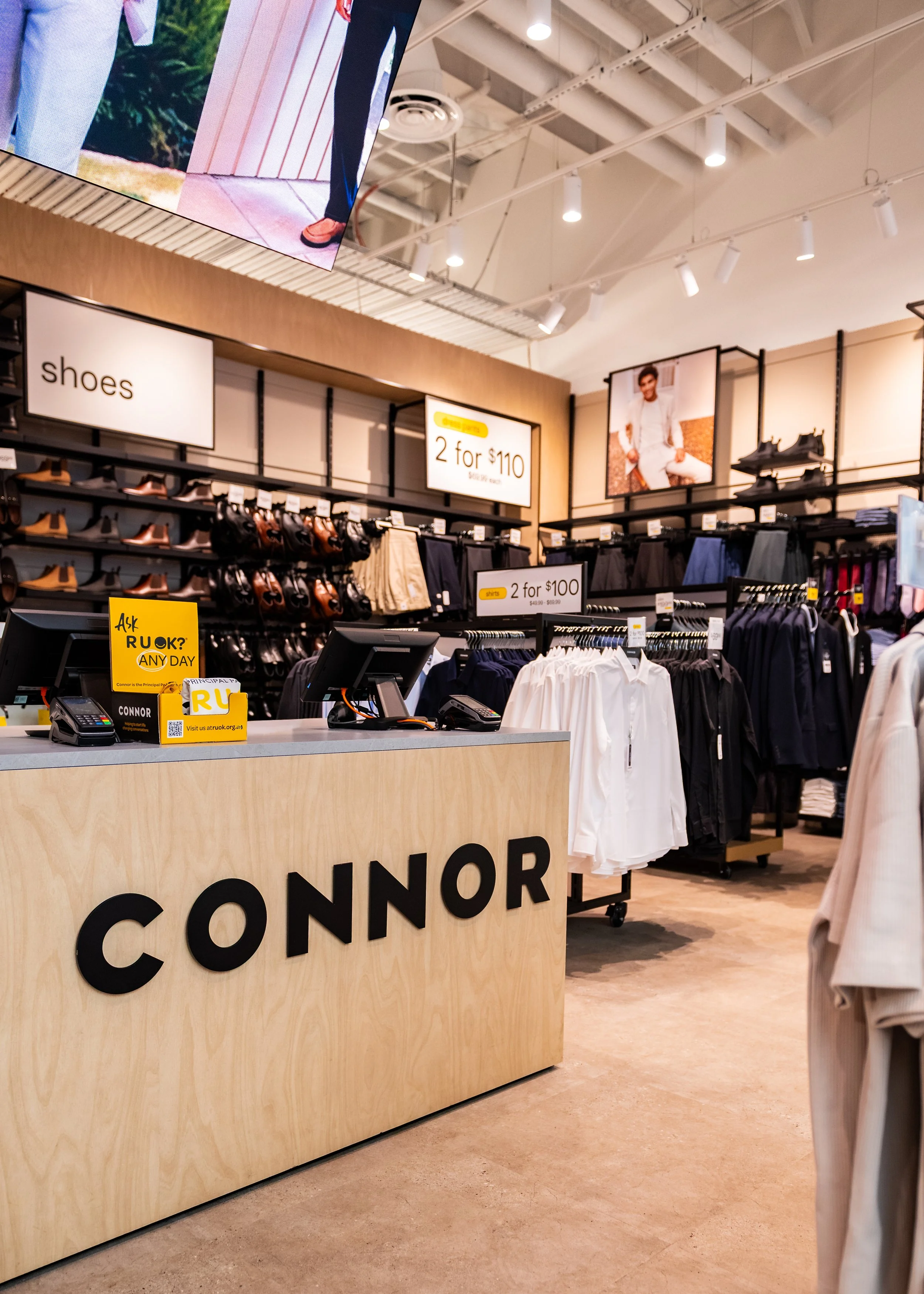 A clothing and shoe store with a wooden cash register counter displaying the brand name 'CONNOR' in large black letters. Behind the counter, shelves display various shoes and pants. There are signs advertising sales, such as '2 for $110' and 'shirts 2 for $100'. The store has bright lighting, and there are large posters of models wearing clothing on the walls.