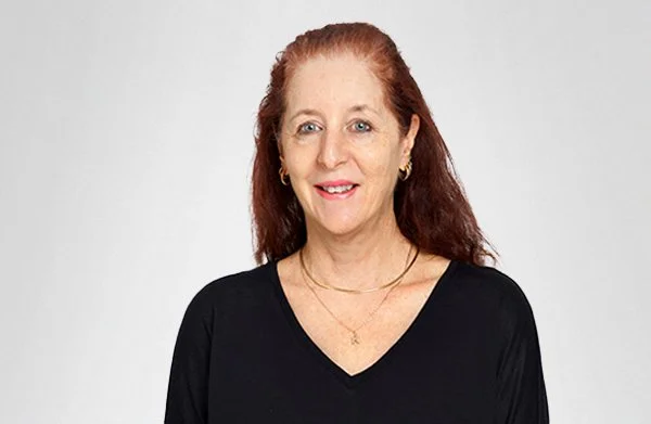 A woman with red hair, wearing a black V-neck top and gold jewelry, smiling against a plain light background.