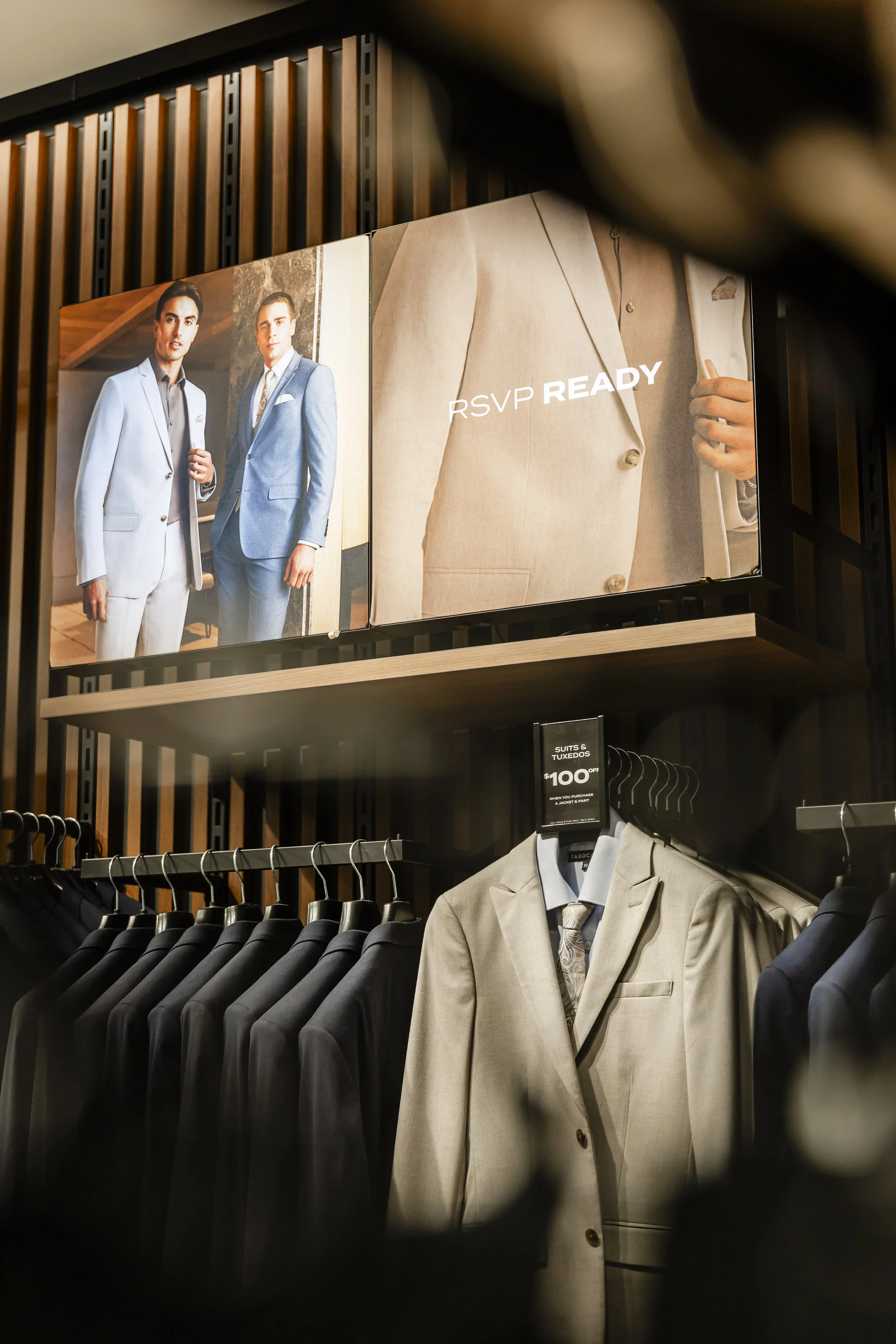 A store display featuring men's suits on racks with a promotional poster for tuxedo rentals above.