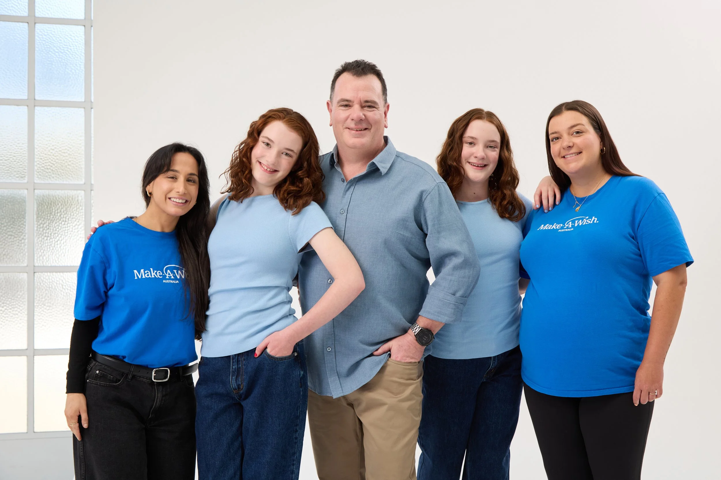 Group of five people standing together, smiling, in a bright room with large windows. Two women are wearing blue shirts with 'Make-A-Wish Australia' written on them, and the others are in casual clothing.