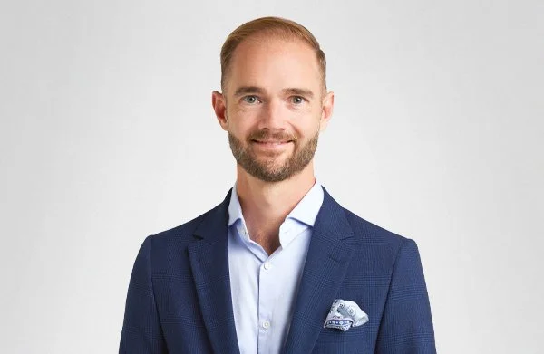 A man in a navy blue suit with a light blue shirt and a patterned pocket square, smiling against a plain white background.