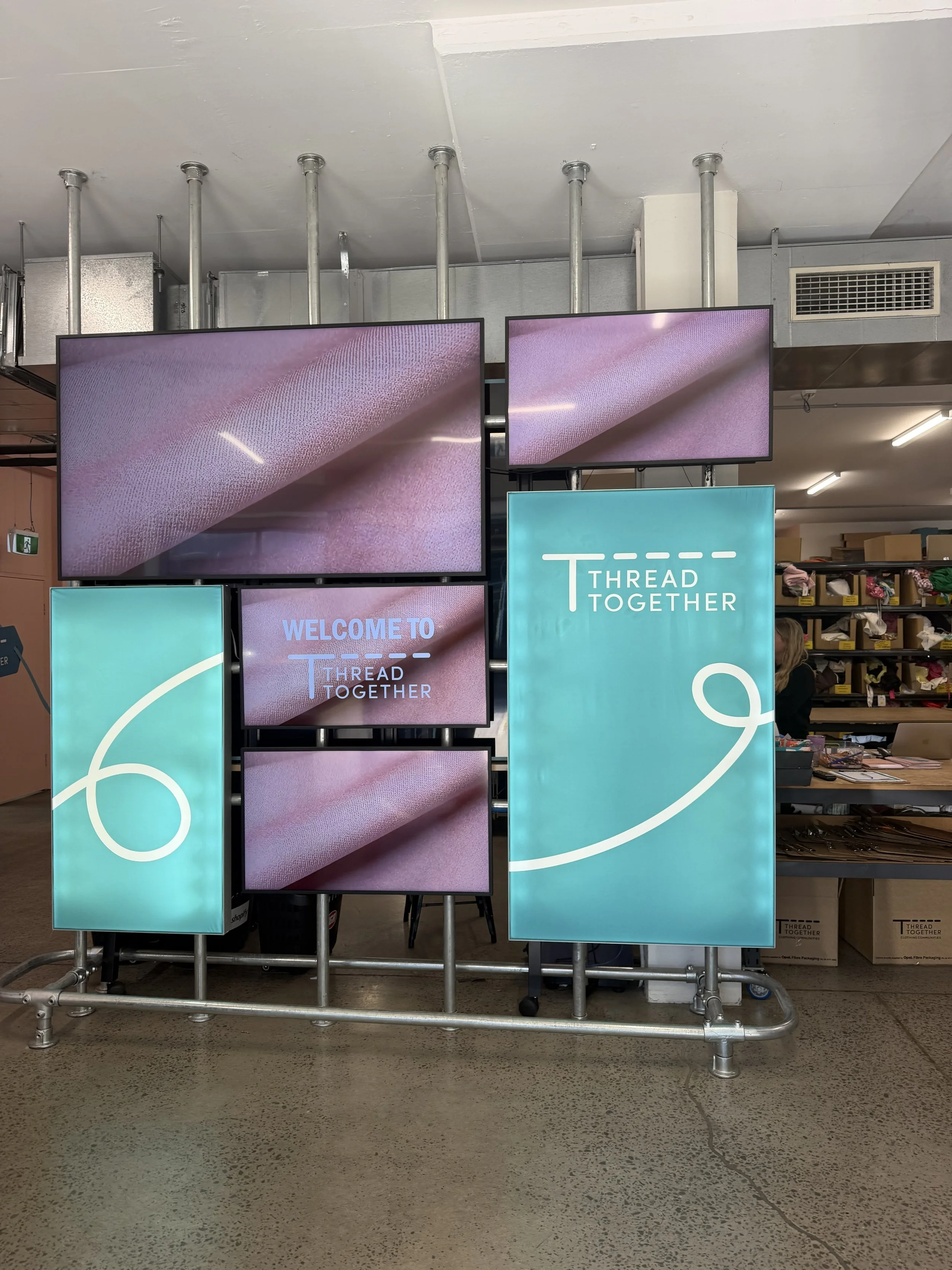Display stand with multiple screens showing the words "Welcome to Thread Together" and a graphic of a white thread loop on a teal background, located in a retail store aisle.
