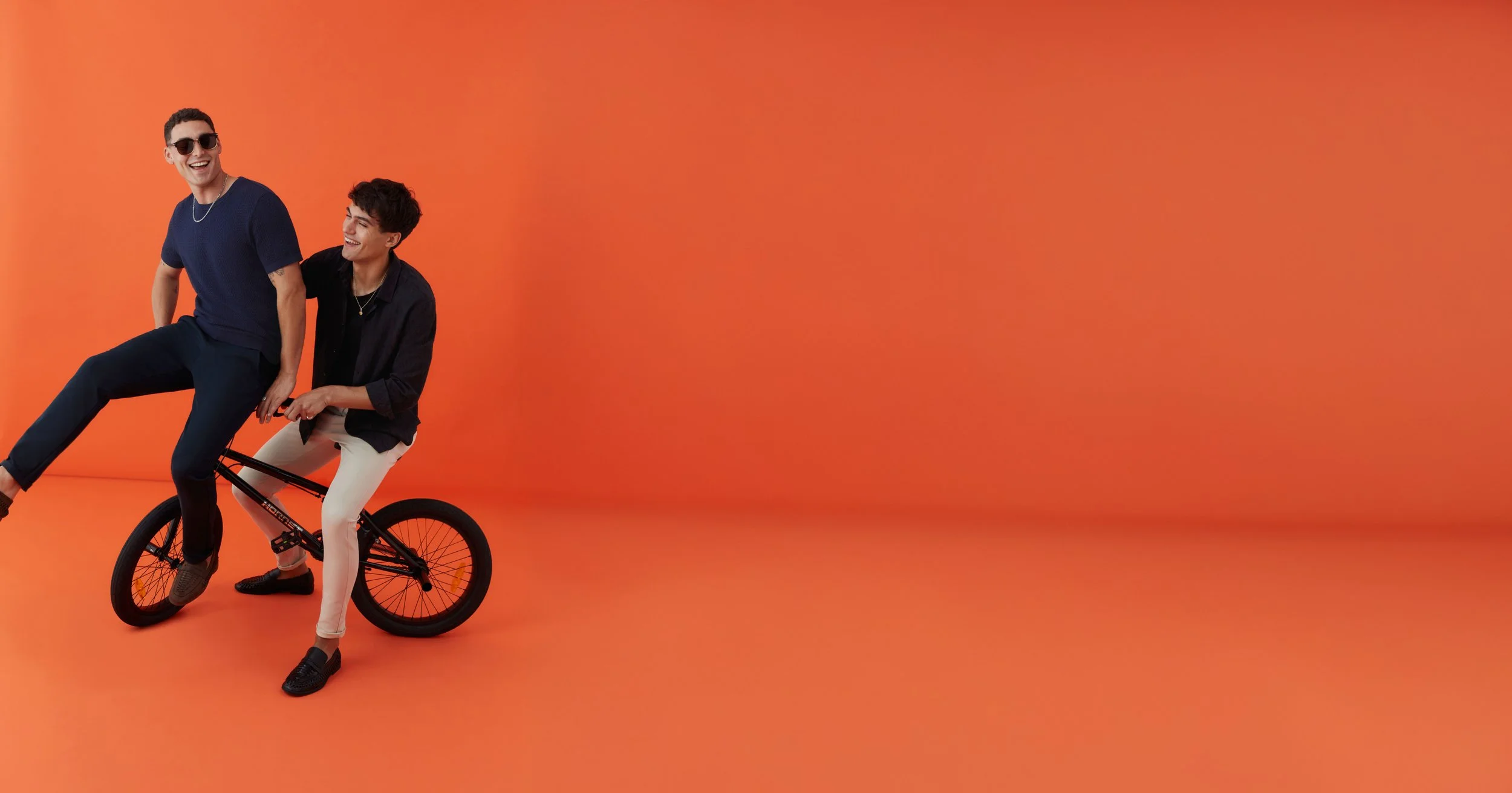 Two young men smiling and laughing, one riding a bicycle while the other sits on the back, against an orange background.