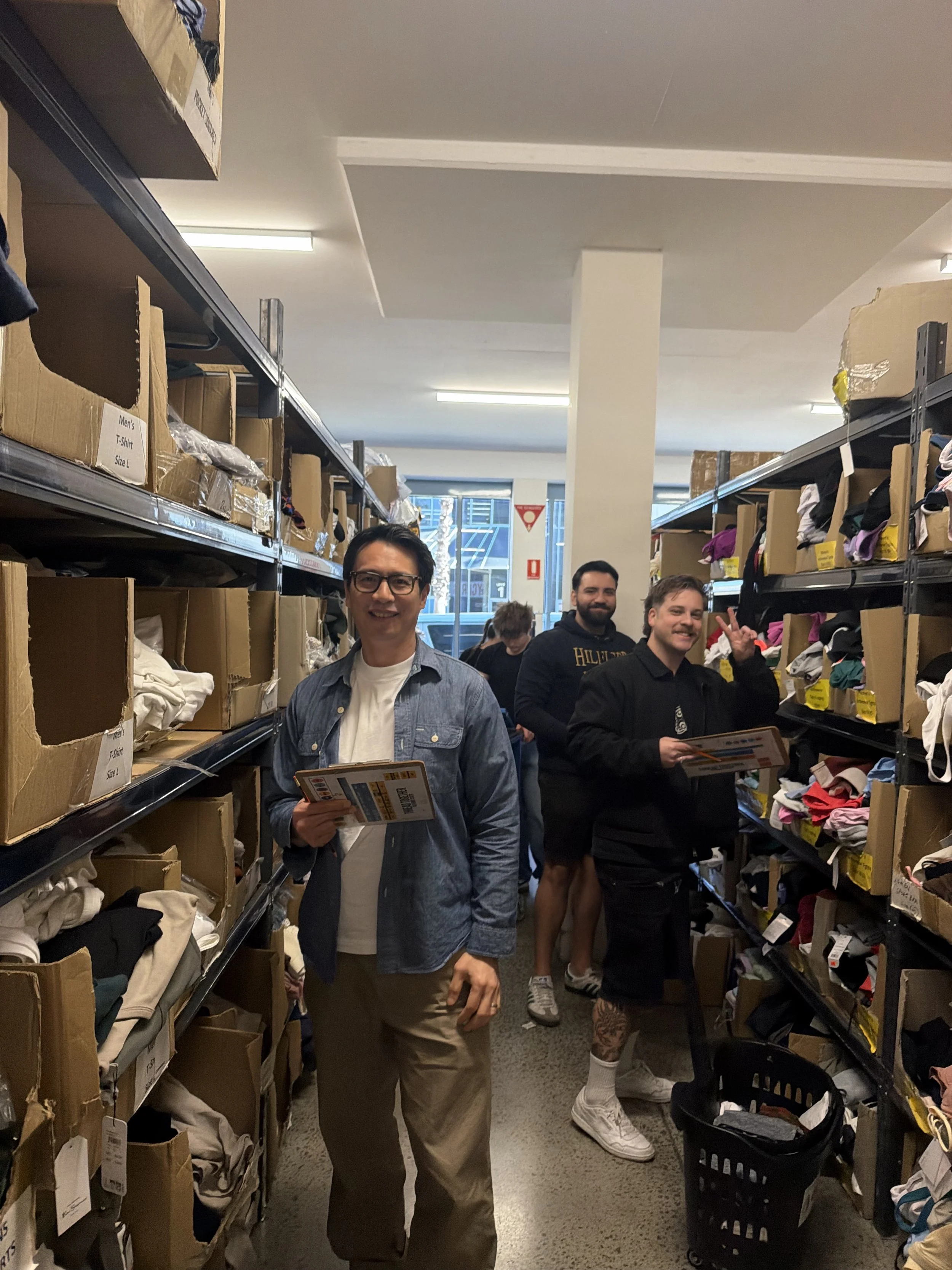 A group of four young men shopping in a thrift store aisle with shelves of folded clothes and shoes, one man in the foreground smiling and holding a tray, two behind him smiling and making peace signs, and one further back browsing.
