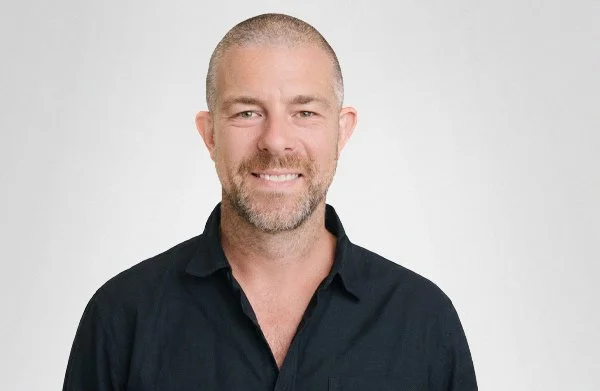 Portrait of a smiling man with a beard, wearing a black button-up shirt, against a plain white background.