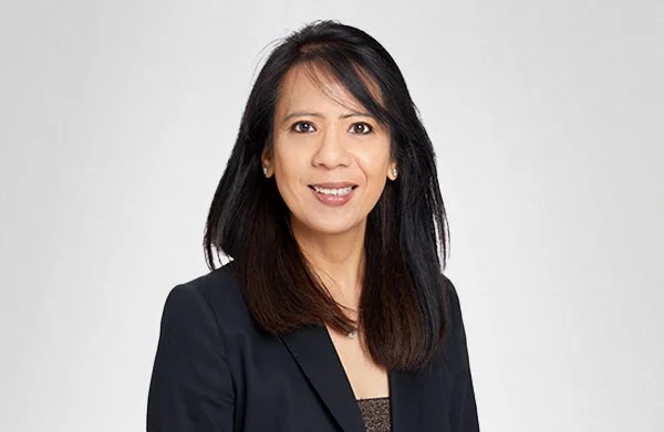 Professional woman with shoulder-length black hair, wearing a black blazer and a dark top, smiling against a plain gray background.