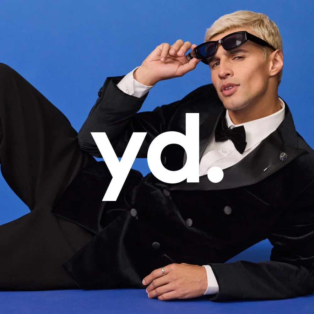 A man dressed in a tuxedo lying on a blue background, holding sunglasses with one hand, with the word 'yd.' written in white across the image.