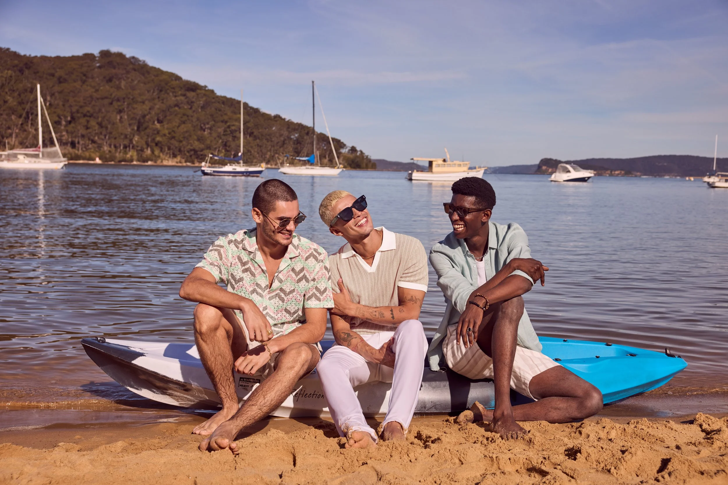 Three friends sitting on a kayak on a sandy beach with boats anchored in the water and a hill in the background under a clear sky.