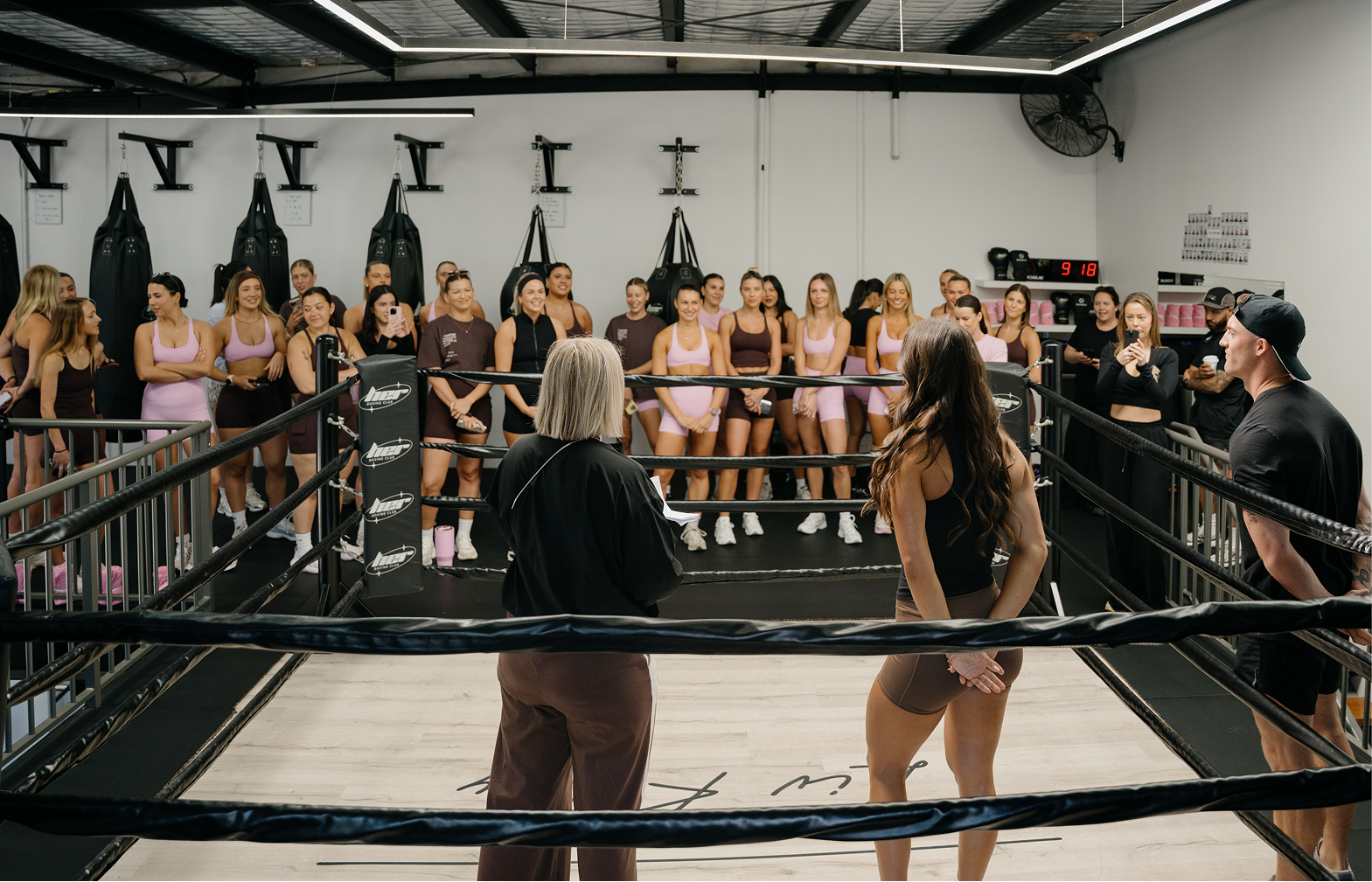 Women in sports bras and shorts standing around a boxing ring in a gym, with an audience watching and taking photos, listening to two women and a man in the ring.