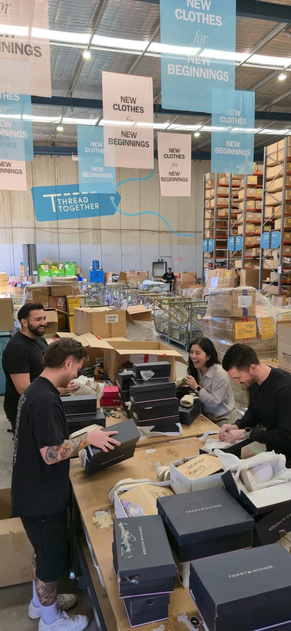 People working at a table in a warehouse, packing boxes labeled 'Women Shoes' with boxes of designer shoes, in a storage area with shelves and banners that read 'New Clothes for New Beginnings' and 'Thread Together'.