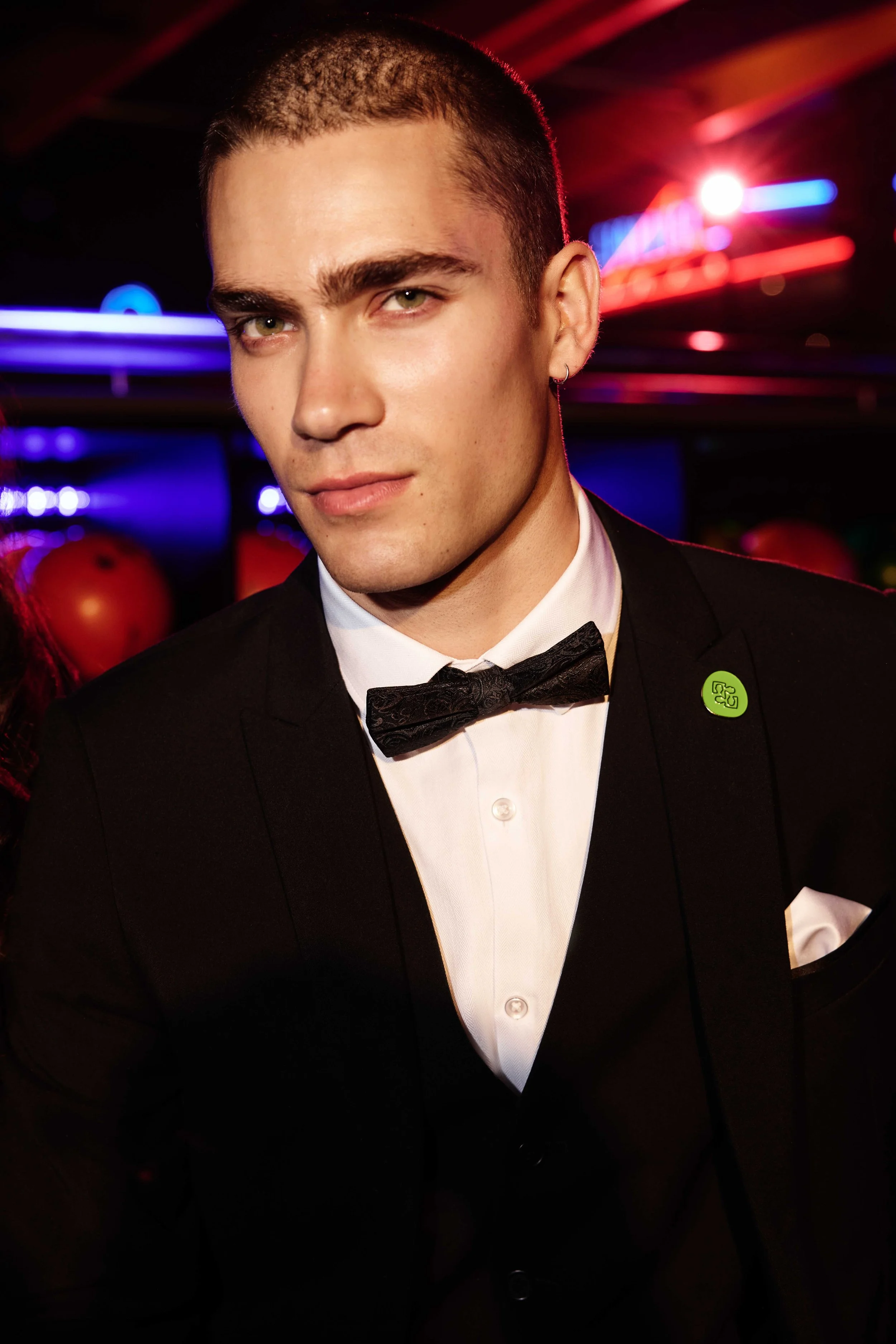 A man in a tuxedo with a bow tie, white shirt, and a green lapel pin against a background with red, blue, and purple neon lights.