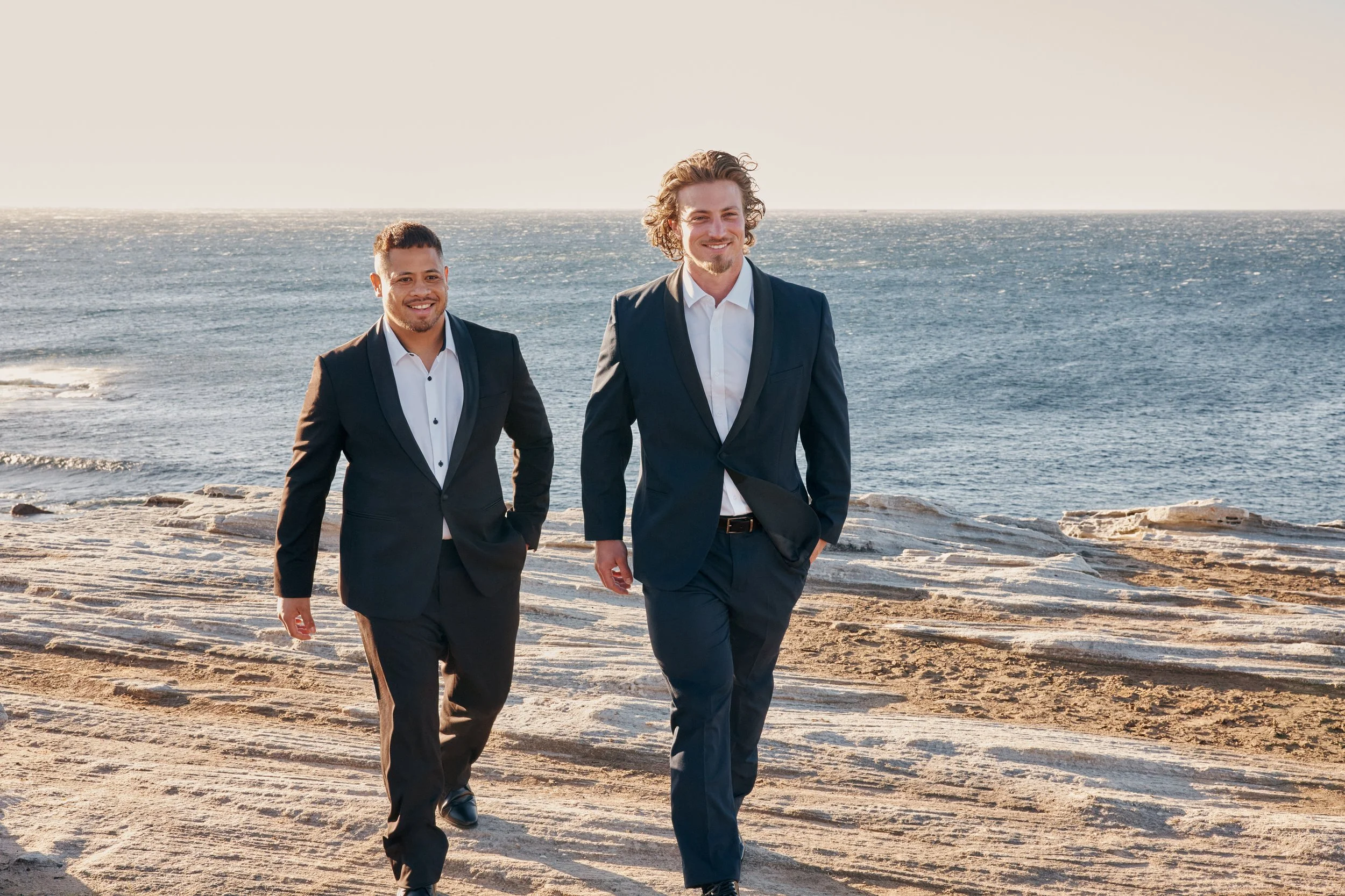 Two men in suits walking along a rocky beach with the ocean in the background, smiling at the camera.