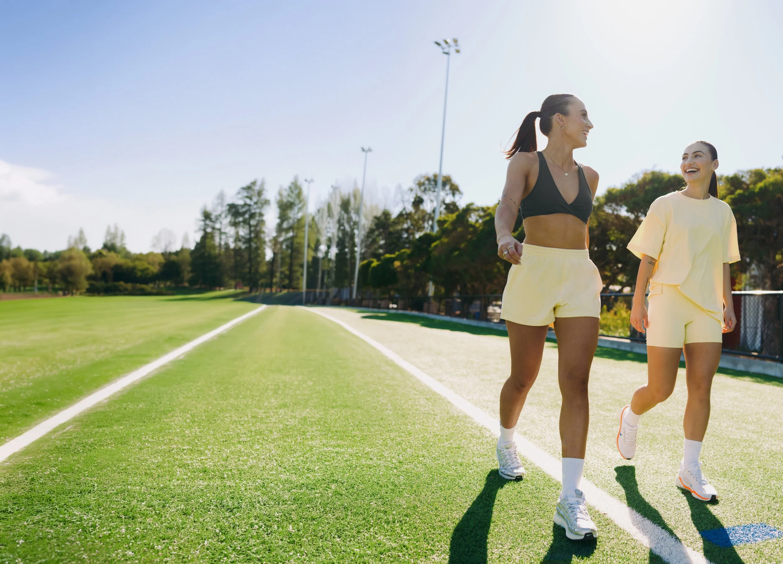 Two women in athletic clothes walking on a track at a sports field, smiling and enjoying a sunny day.