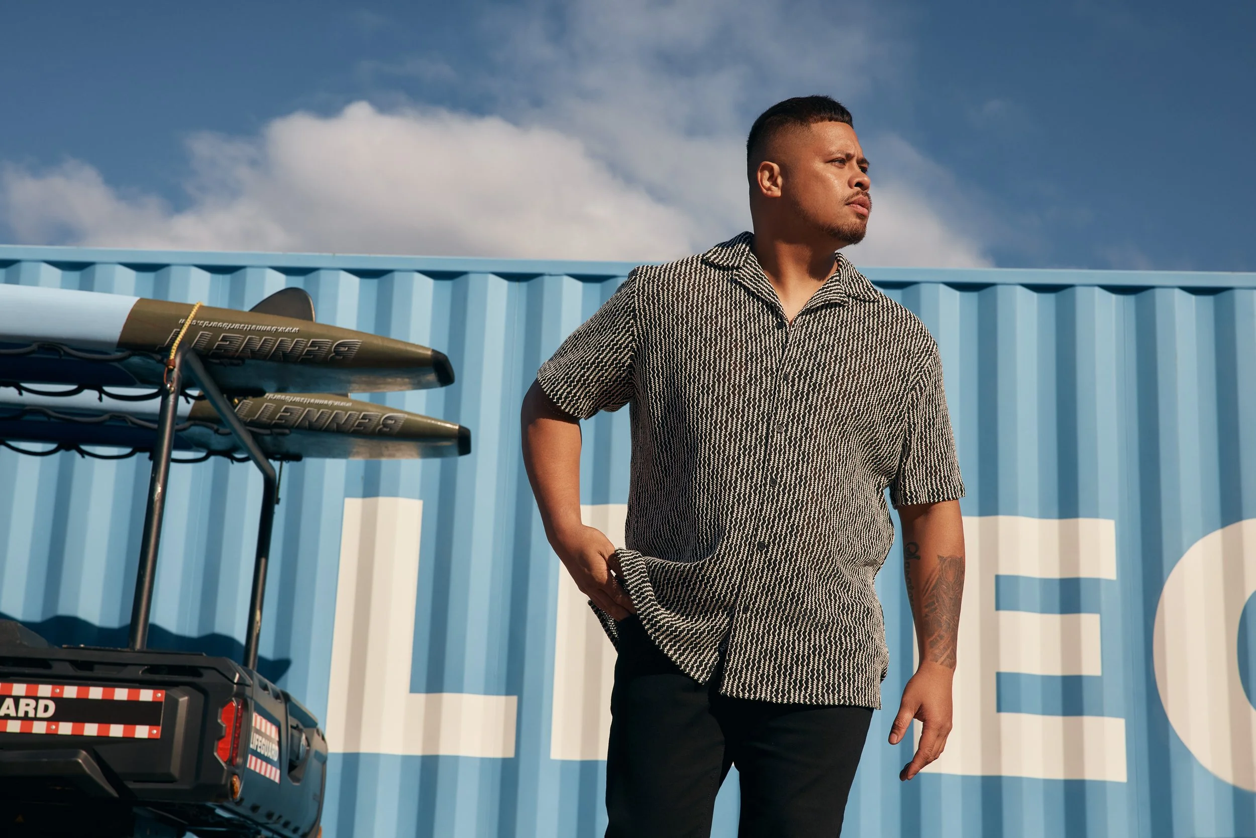 A man with short haircut and a beard, wearing a patterned short-sleeve shirt, stands in front of a blue wall with clouds visible in the sky. There are folded chairs and a portable generator with an emergency sticker nearby.