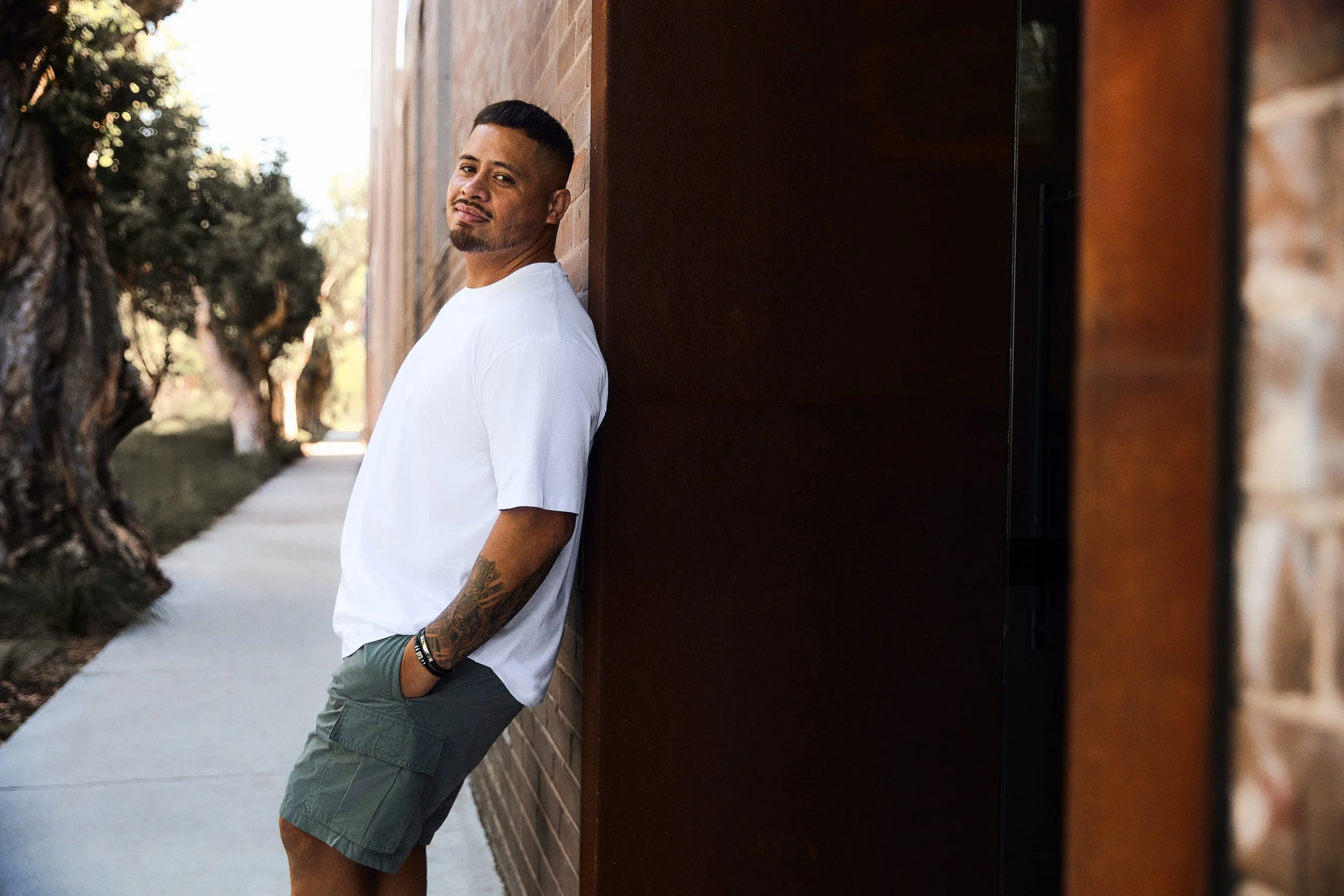 A man with tattoos on his arm leaning against a dark brown wall outdoors, smiling slightly, with trees and a sidewalk in the background.