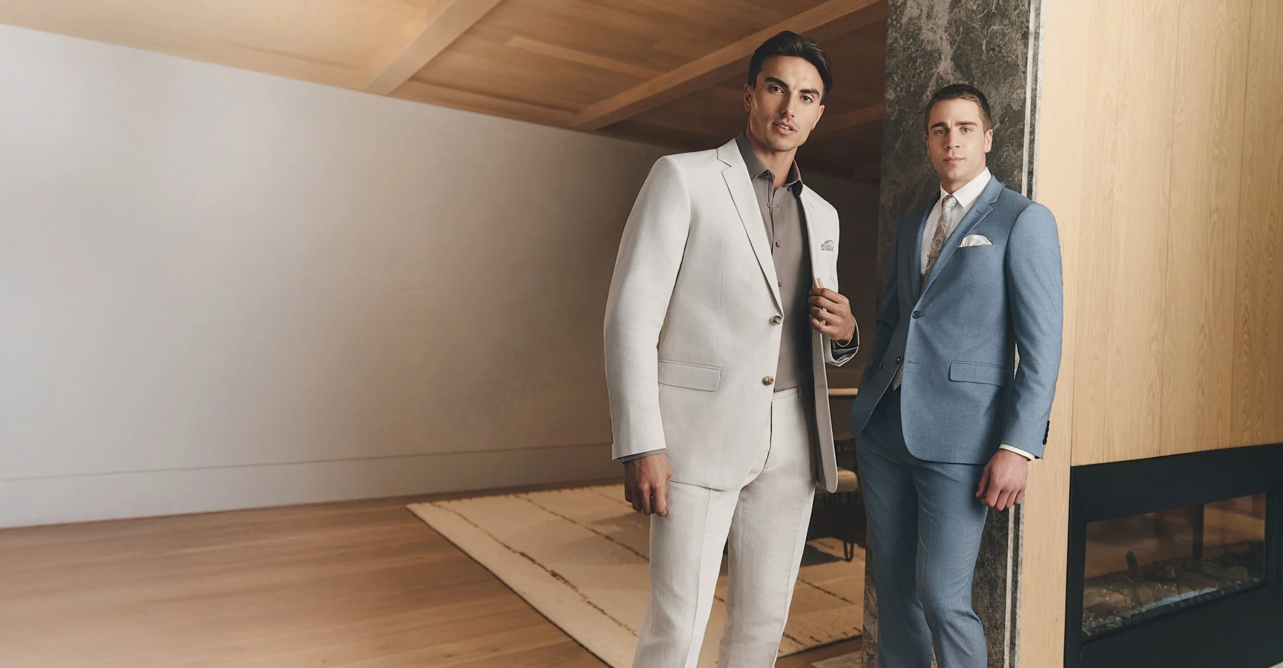 Two men wearing suits standing in a modern interior space. The man on the left wears a cream-colored suit with a dark shirt underneath, and the man on the right wears a light blue suit with a white shirt and patterned tie. They are standing near a wall with wood paneling and a stone accent element.