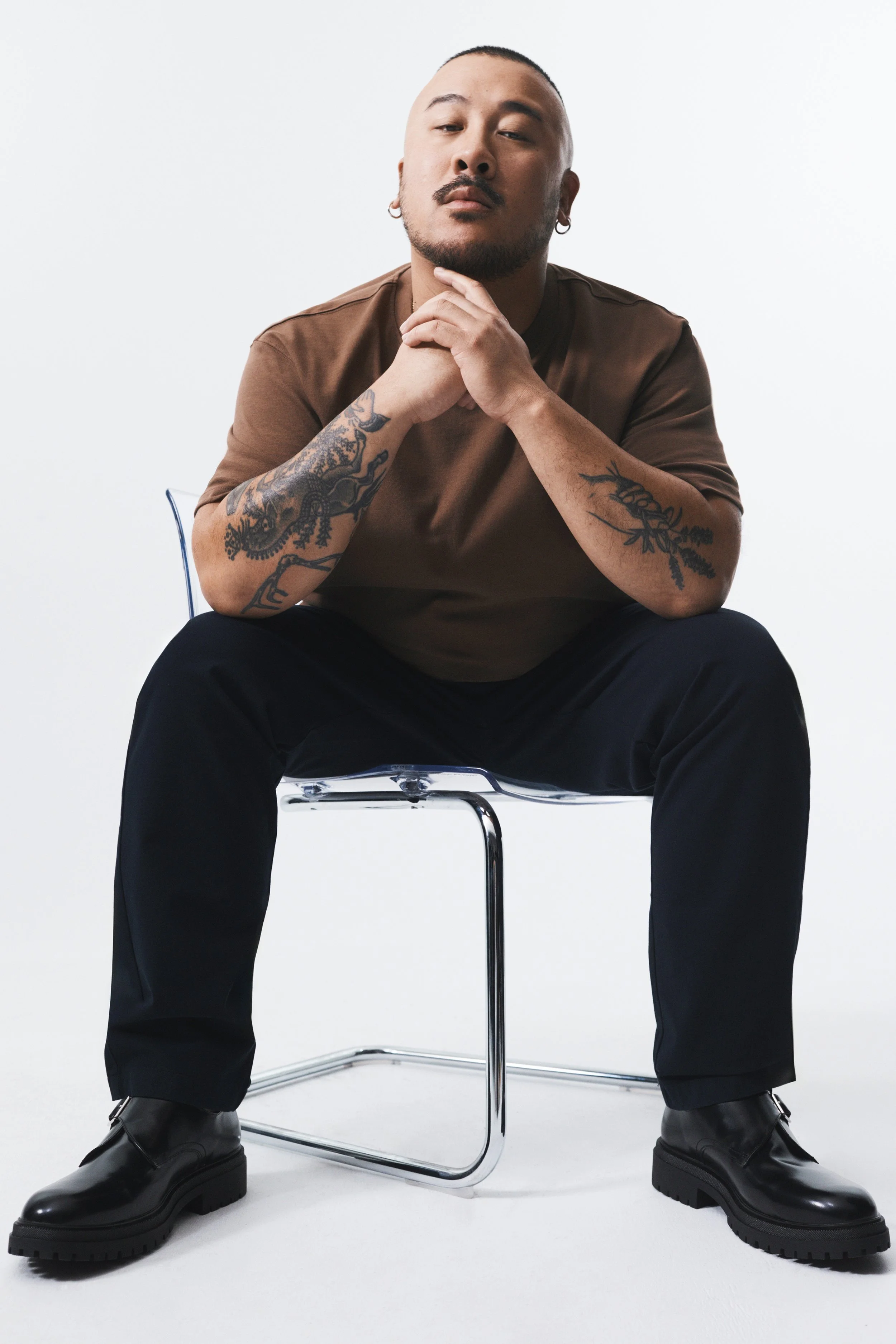 A man with tattoos on both arms sitting on a clear plastic chair, wearing a brown t-shirt, black pants, black boots, and earrings, posing against a plain white background.