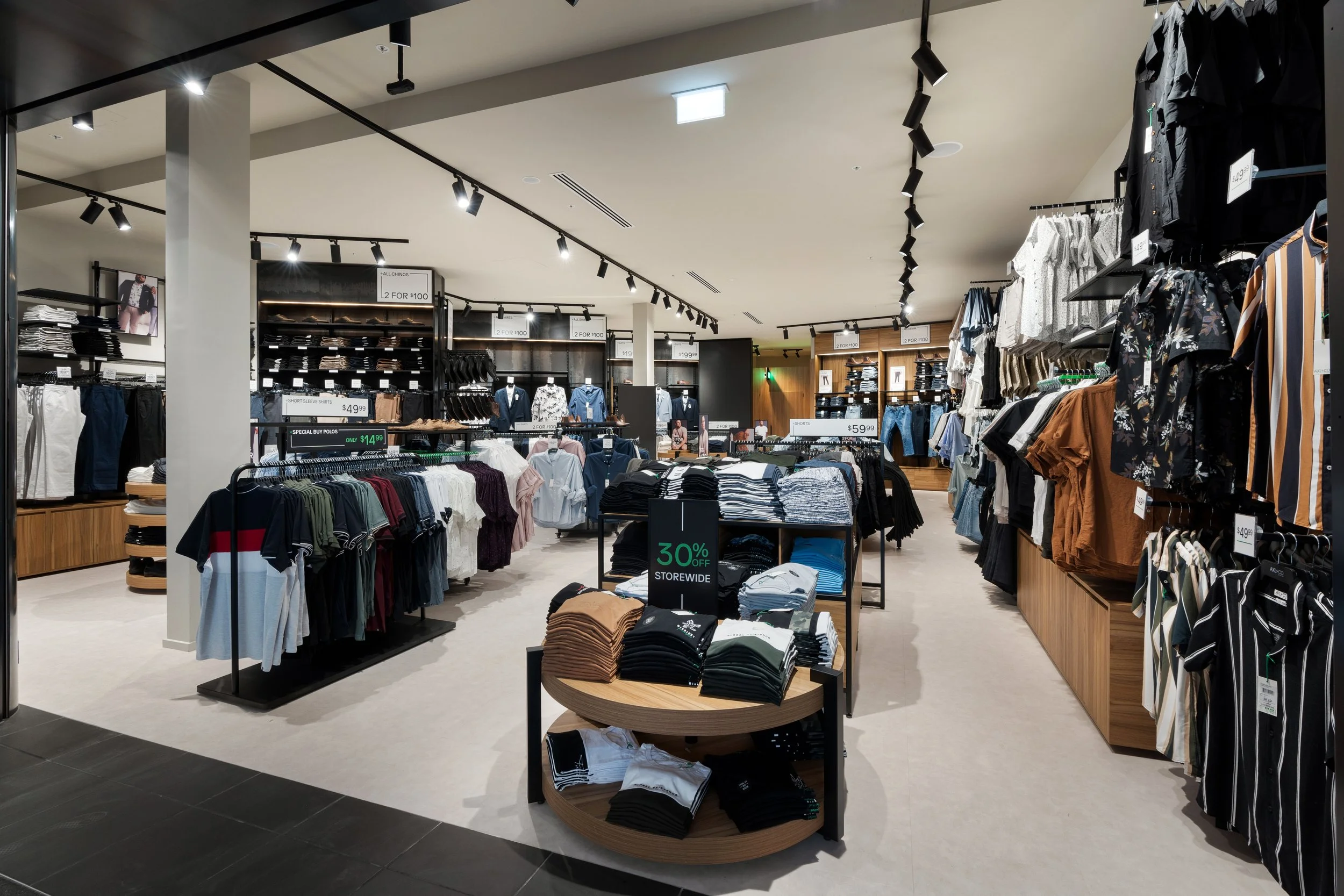 Interior of a clothing store with various racks and shelves of men's and women's apparel, including shirts, pants, and jackets, with sale signs indicating discounts.