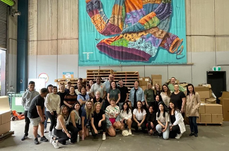 Group of people standing together in a warehouse, with large colorful fabric artwork hanging on the wall behind them and stacks of cardboard boxes nearby.
