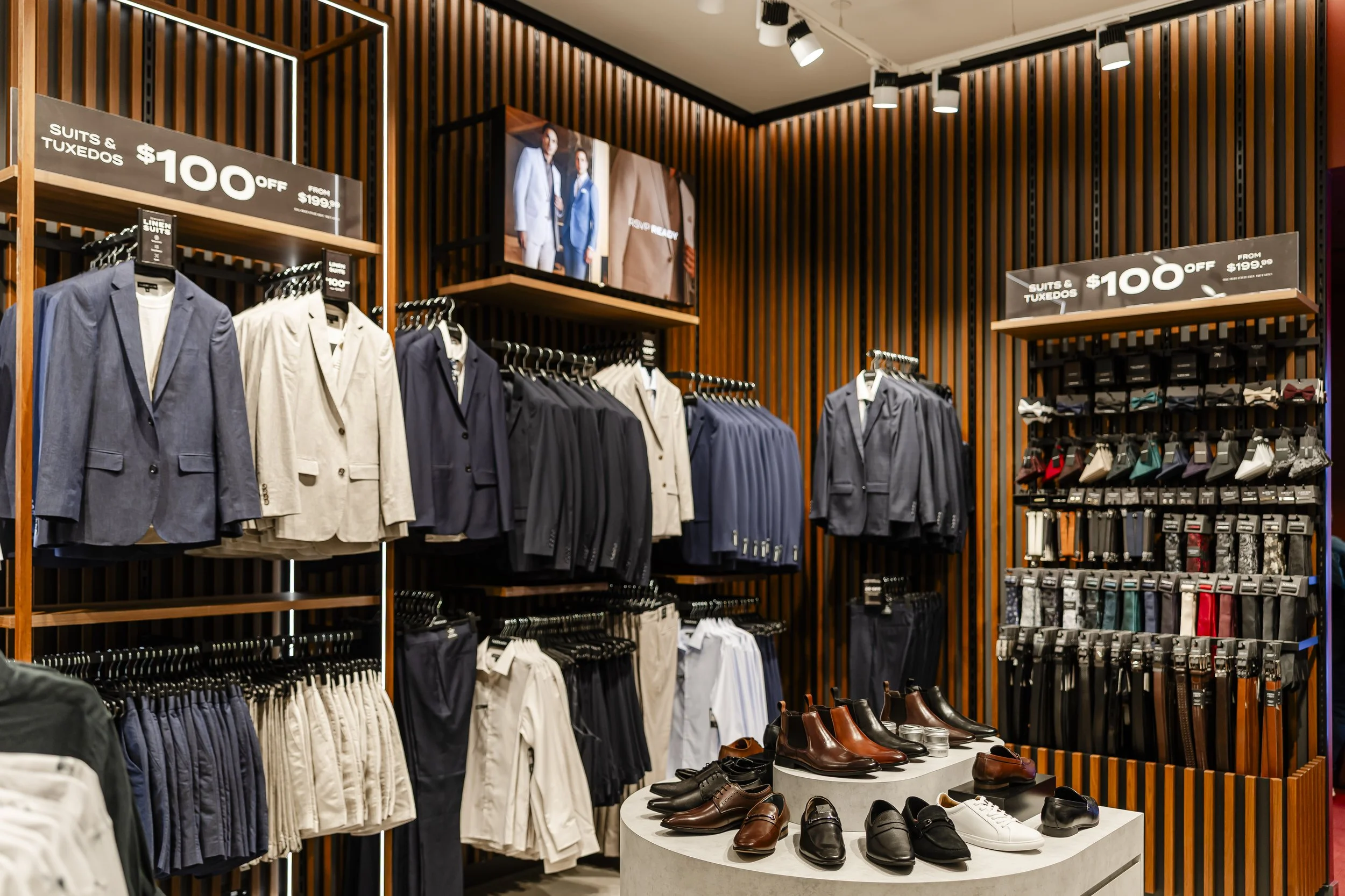 Men's suits and tuxedos on hangers and shoes on a round display table inside a clothing store.