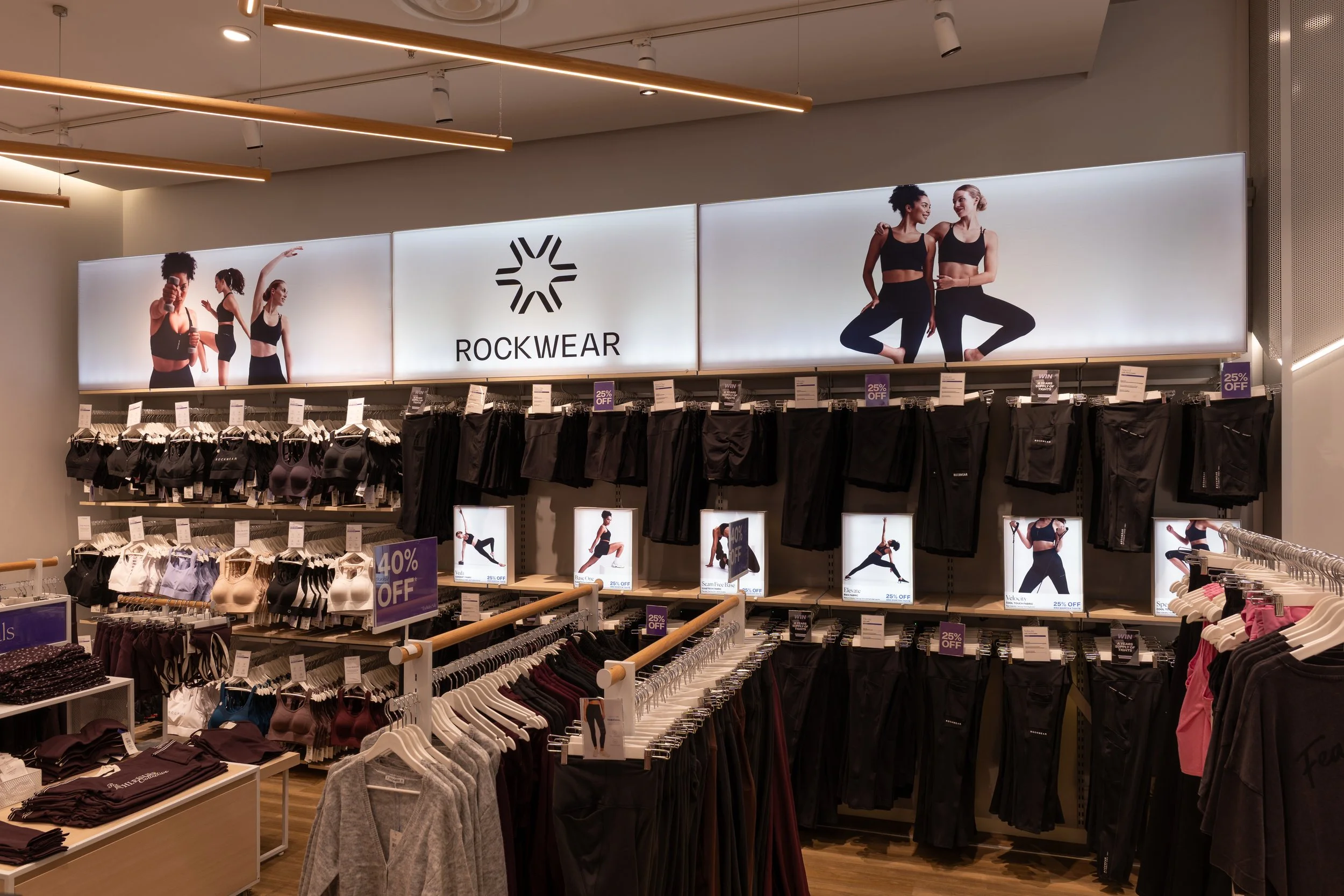 Inside a retail clothing store, display advertising for Rockwear activewear featuring two women in workout clothes, with racks of clothing below and promotional signs offering discounts.
