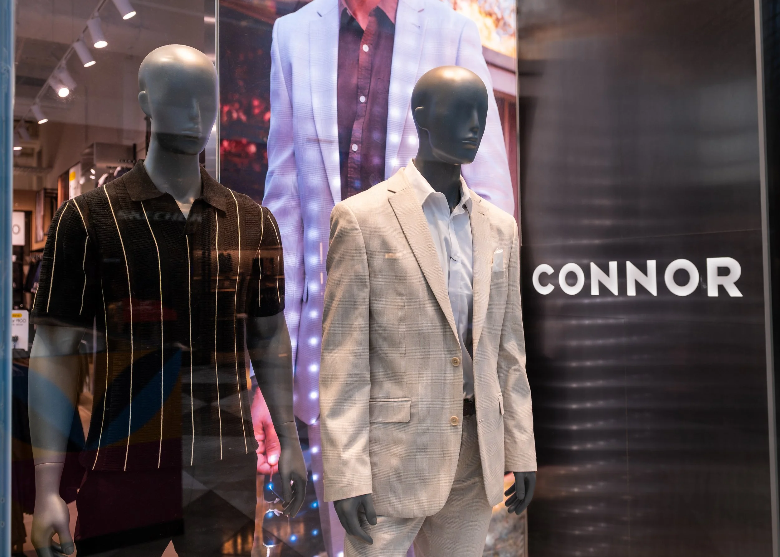 Store display window featuring three mannequins dressed in men's clothing. The left mannequin wears a dark, short-sleeved polo shirt with white and red vertical stripes. The right mannequin wears a light-colored, plaid suit with a matching blazer and shorts, and a white dress shirt underneath. Behind the mannequins, there is a large poster of a man in a pink and white suit jacket with a dark shirt, and the brand name 'CONNOR' is partially visible at the bottom right.
