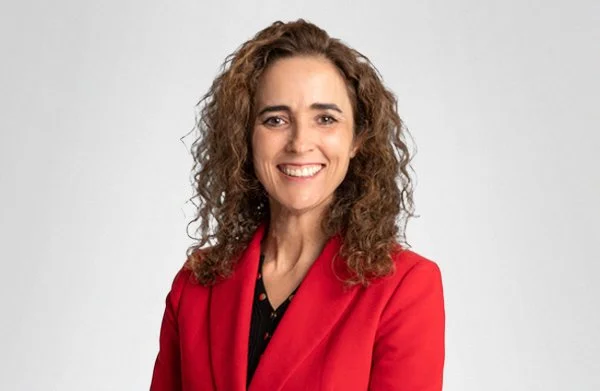 A woman with curly brown hair wearing a red blazer and black top, smiling against a light gray background.