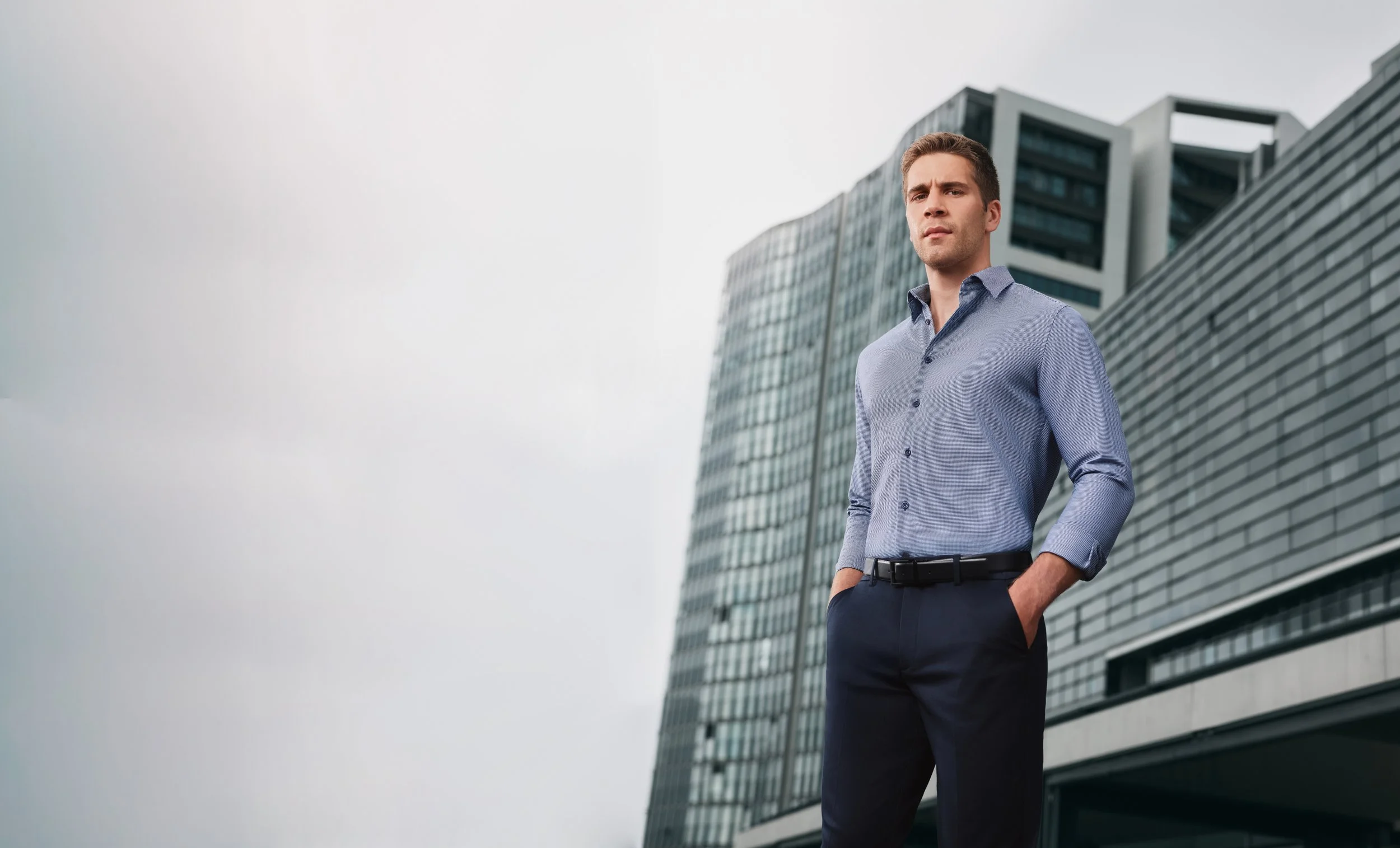A young man in business attire stands outdoors in front of modern office buildings, looking confidently at the camera with hands in his pockets.