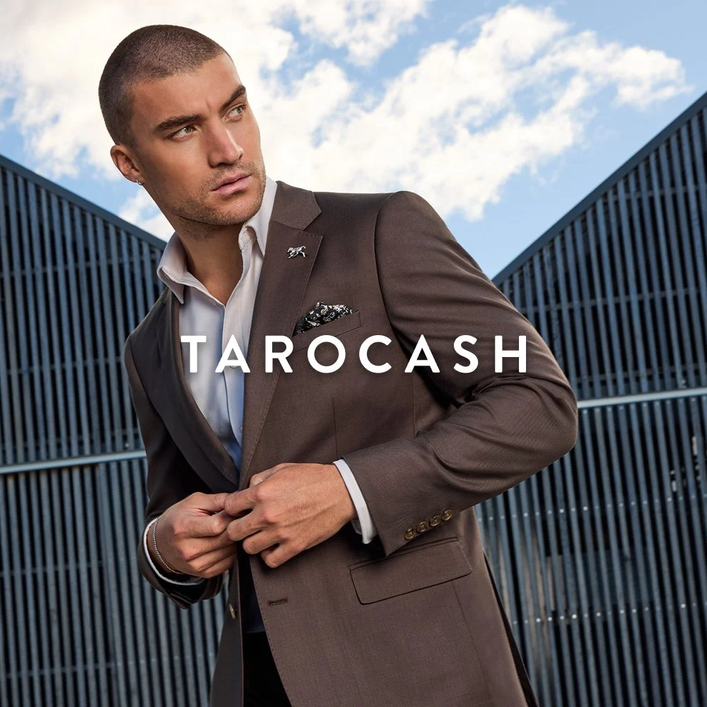 A man in a brown suit and white dress shirt stands outdoors in front of modern buildings with a cloudy sky. He has short hair, is adjusting his suit jacket, and looks to the side. The word 'TAROCASH' is written across the image.