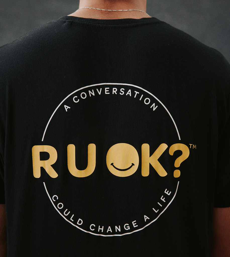 Person wearing a black t-shirt with the text 'A CONVERSATION COULD CHANGE A LIFE' and a smiley face incorporated into the word 'R U OK?'