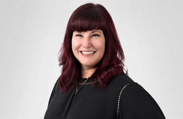 A woman with shoulder-length dark red hair, smiling, wearing a black top with silver stitching details, standing against a plain white background.