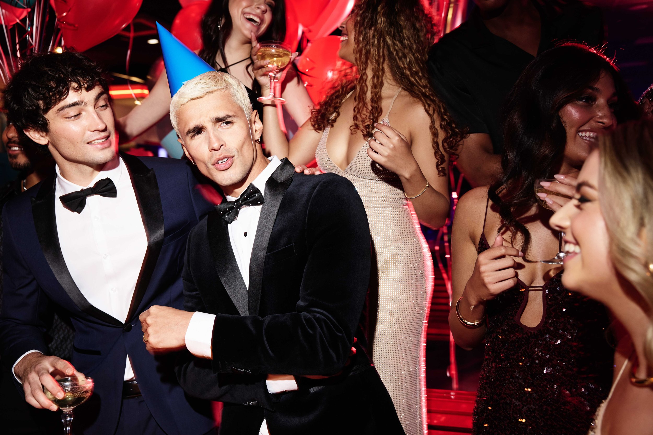 People at a party wearing formal attire with women in dresses and men in tuxedos, celebrating with balloons and drinks, some wearing festive party hats and leis, in a lively setting with colorful lighting.