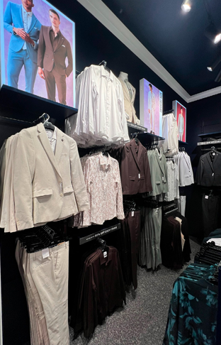 Clothing display in a retail store featuring jackets, blouses, and pants with photographs of models on digital screens above.