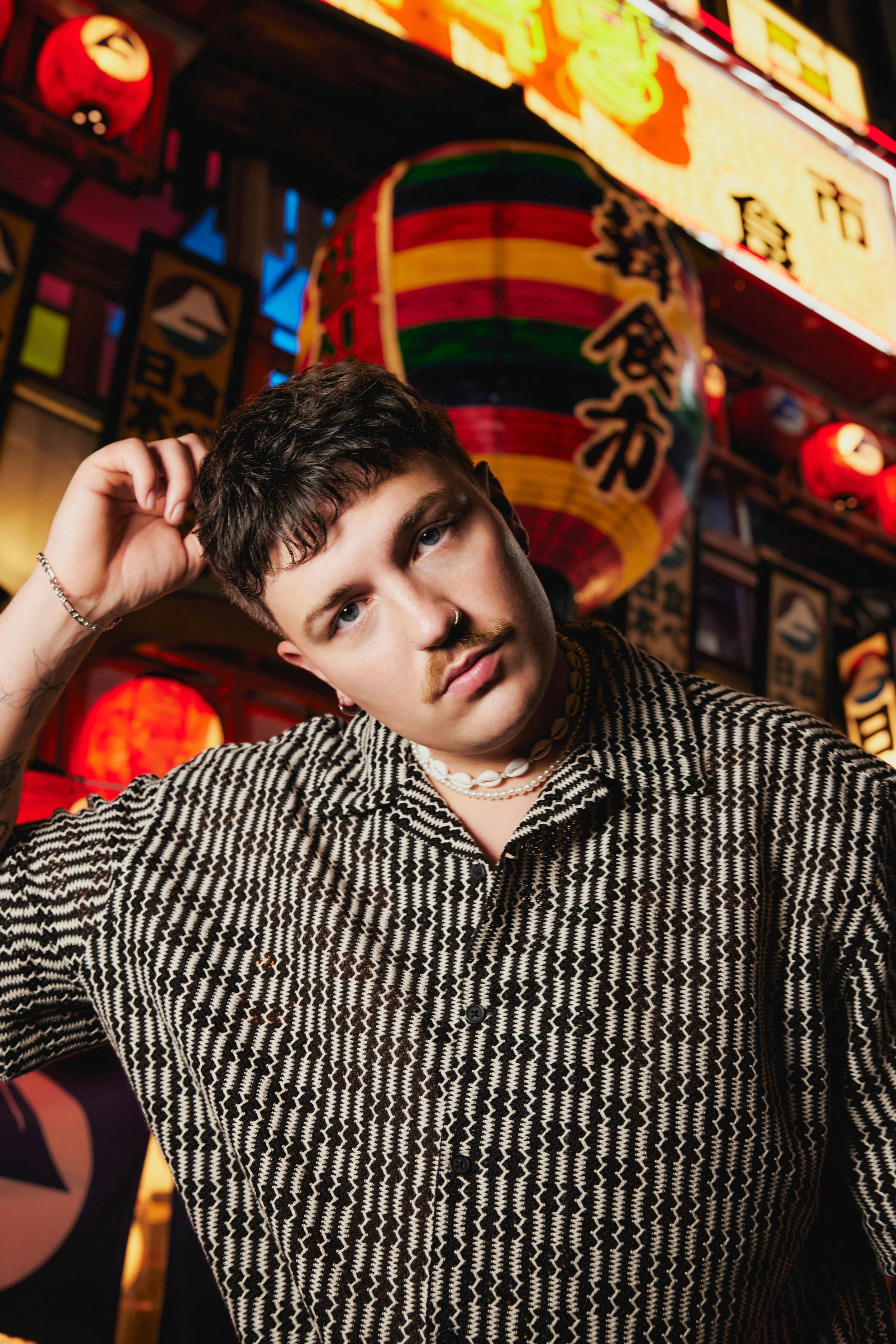 A young person with short, dark, curly hair, wearing a patterned shirt and white shell necklace, is posing with one hand on their head in a vibrant, decorated location with Japanese lanterns and bright signs.