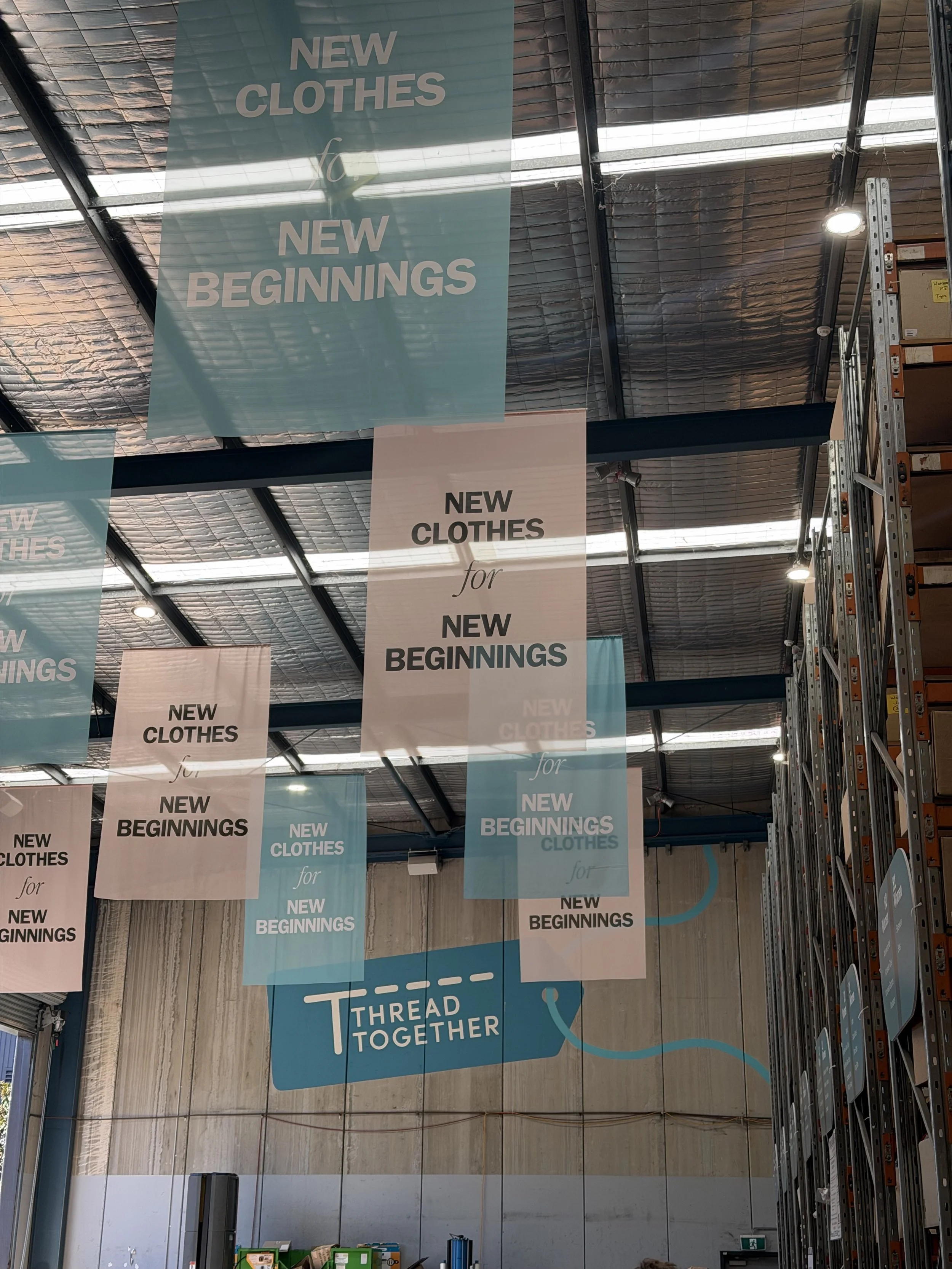 Interior of a warehouse or store with banners hanging from the ceiling that read, 'NEW CLOTHES for NEW BEGINNINGS' and 'THREAD TOGETHER'.