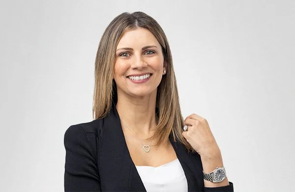 A woman with shoulder-length brown hair smiling, wearing a black blazer, white top, a necklace with a heart pendant, a watch, and a ring, posing against a gray background.