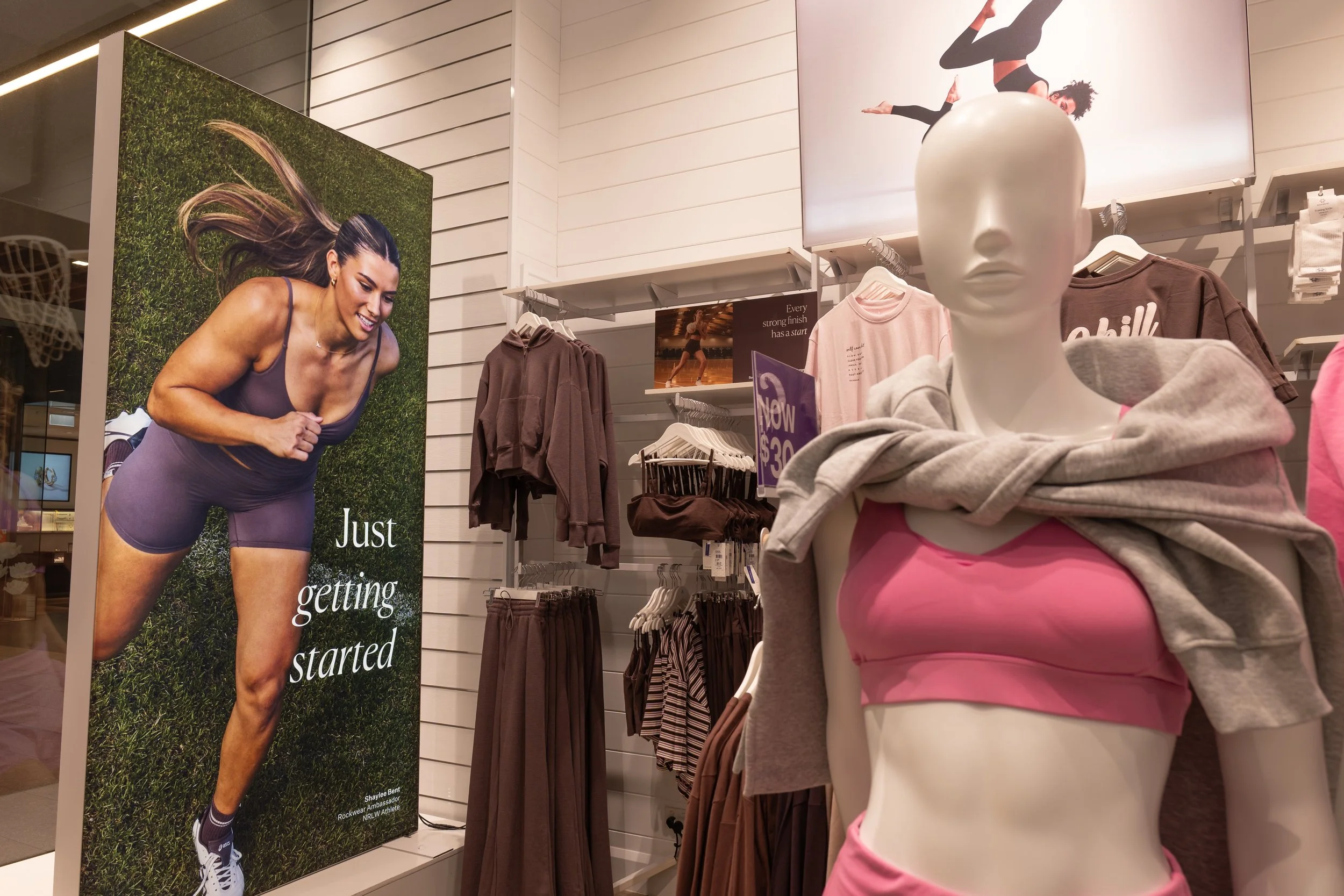 A retail store display featuring athletic clothing and a mannequin dressed in a pink sports bra and gray hoodie, with a large poster of a woman in workout attire running on a grassy field.
