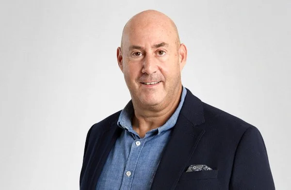 Headshot of man with a bald head, wearing a navy blazer over a blue button-up shirt, smiling against a plain light gray background.