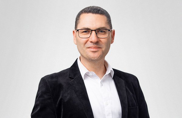 Headshot of man with glasses, wearing a white shirt and black blazer, smiling against a plain light gray background.
