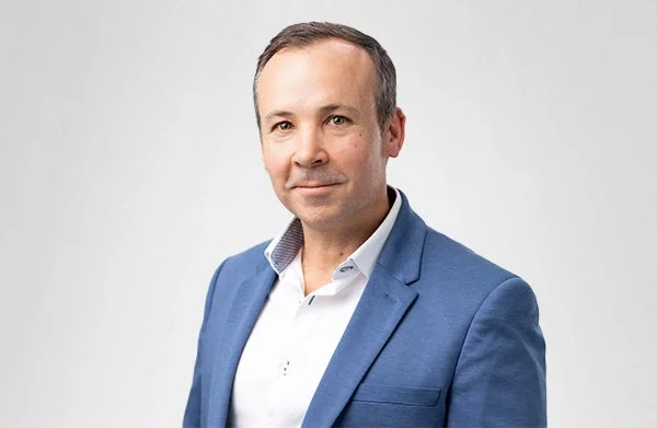 Headshot of man in a blue blazer and white shirt against a plain background.