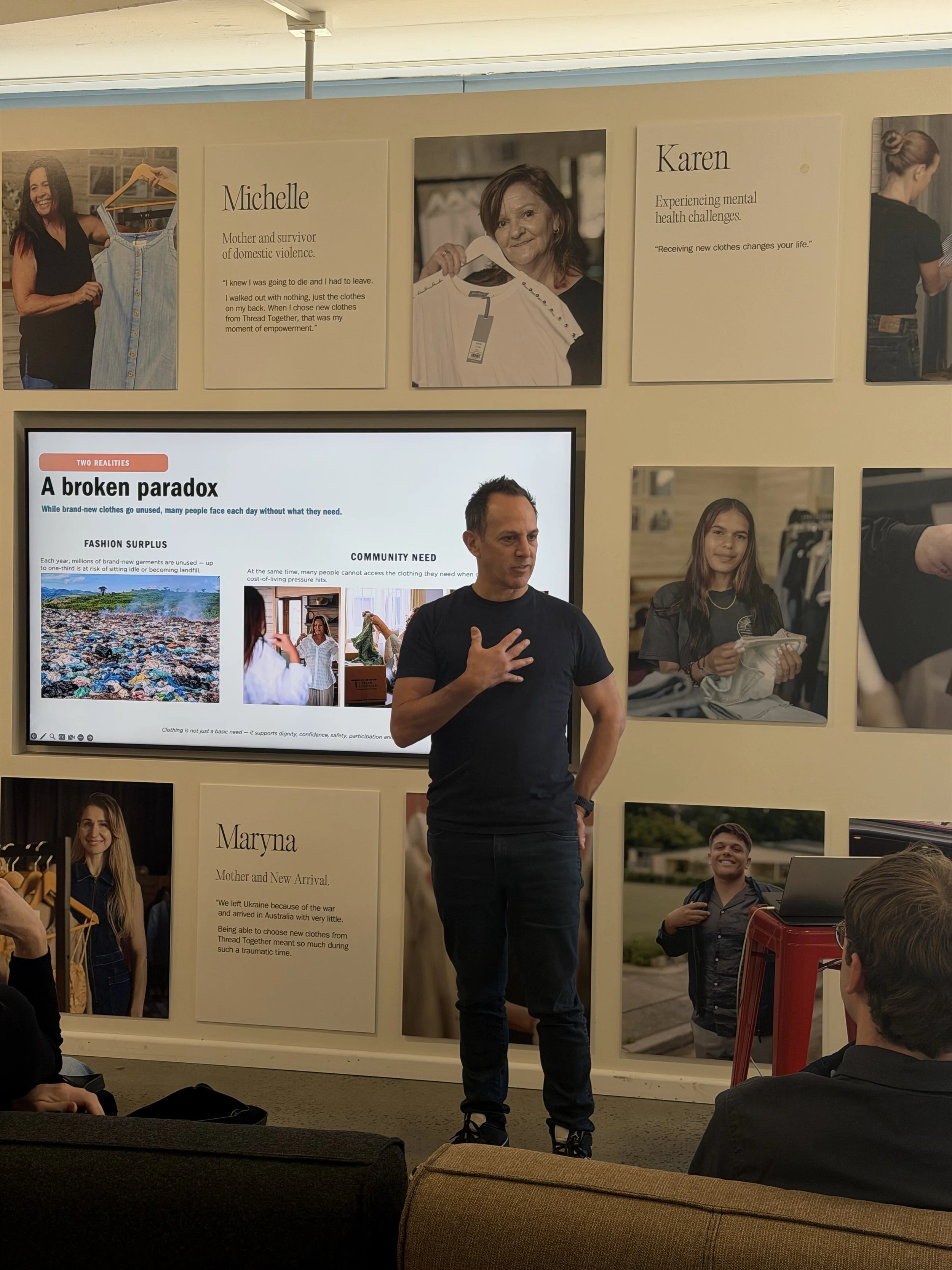 A man stands and speaks to an audience in front of a display wall with various portraits and informational posters about different individuals. The setting appears to be an indoor event or presentation.