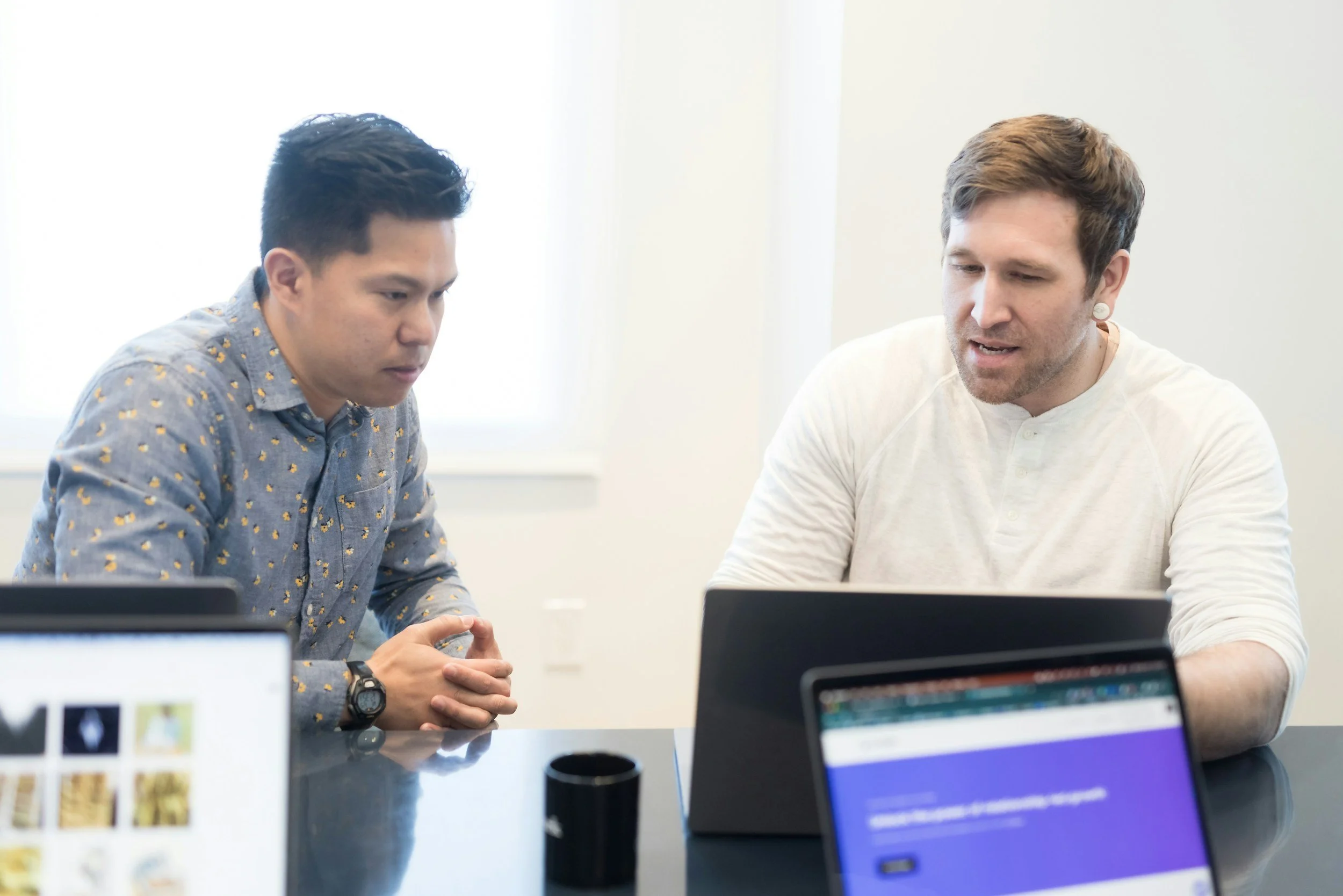 Two men seated at a meeting table engaged in conversation, with laptops and computer screens in front of them in a bright office setting.
