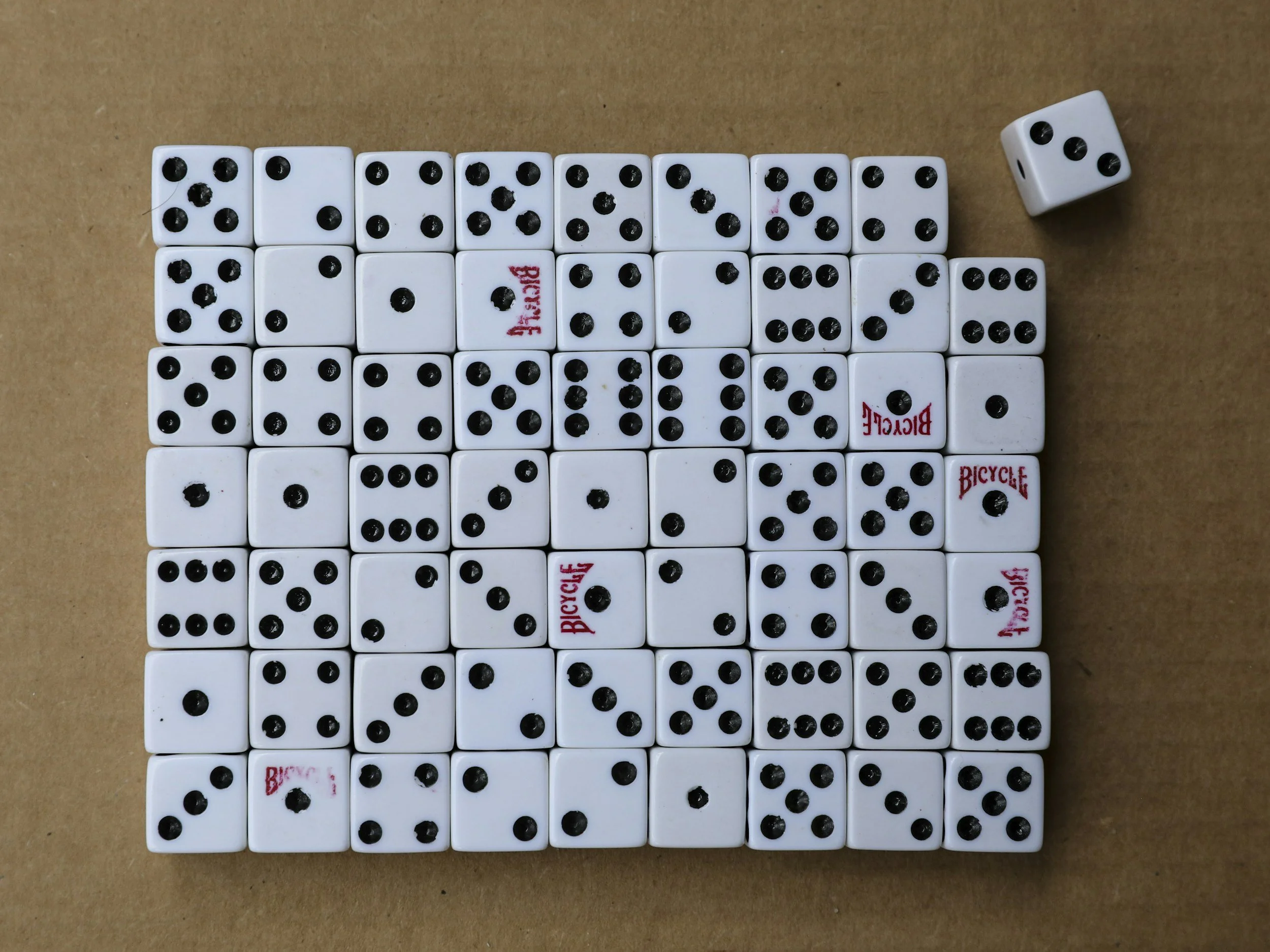 A collection of 36 white dice with black dots, arranged in a grid on a beige surface, with two dice showing the numbers 1 and 6, and some dice with the brand name 'BICYCLE' visible.