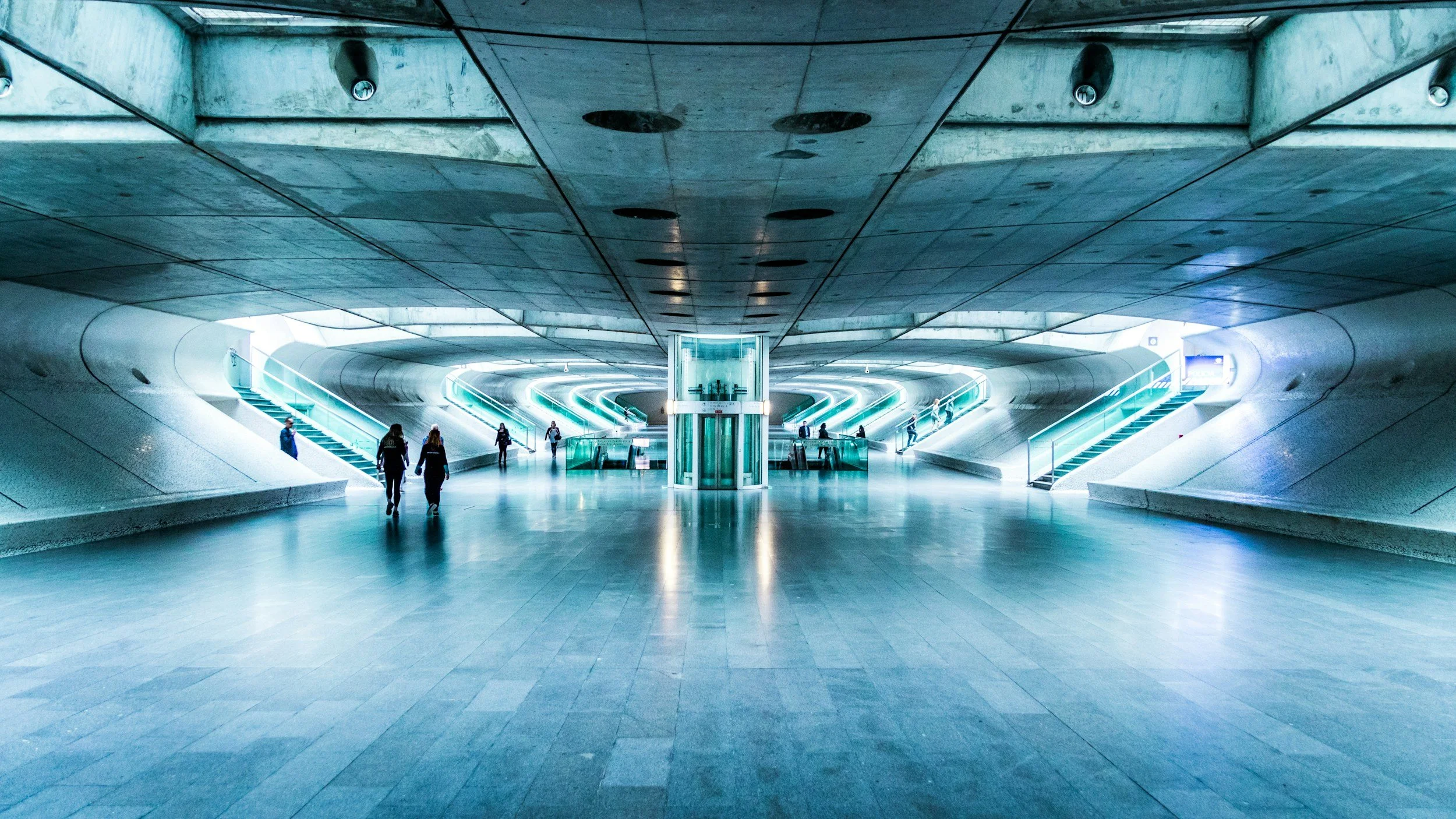 Large modern underground subway station with curved white walls, stairs, and escalators leading to different levels, with a few people walking and standing.