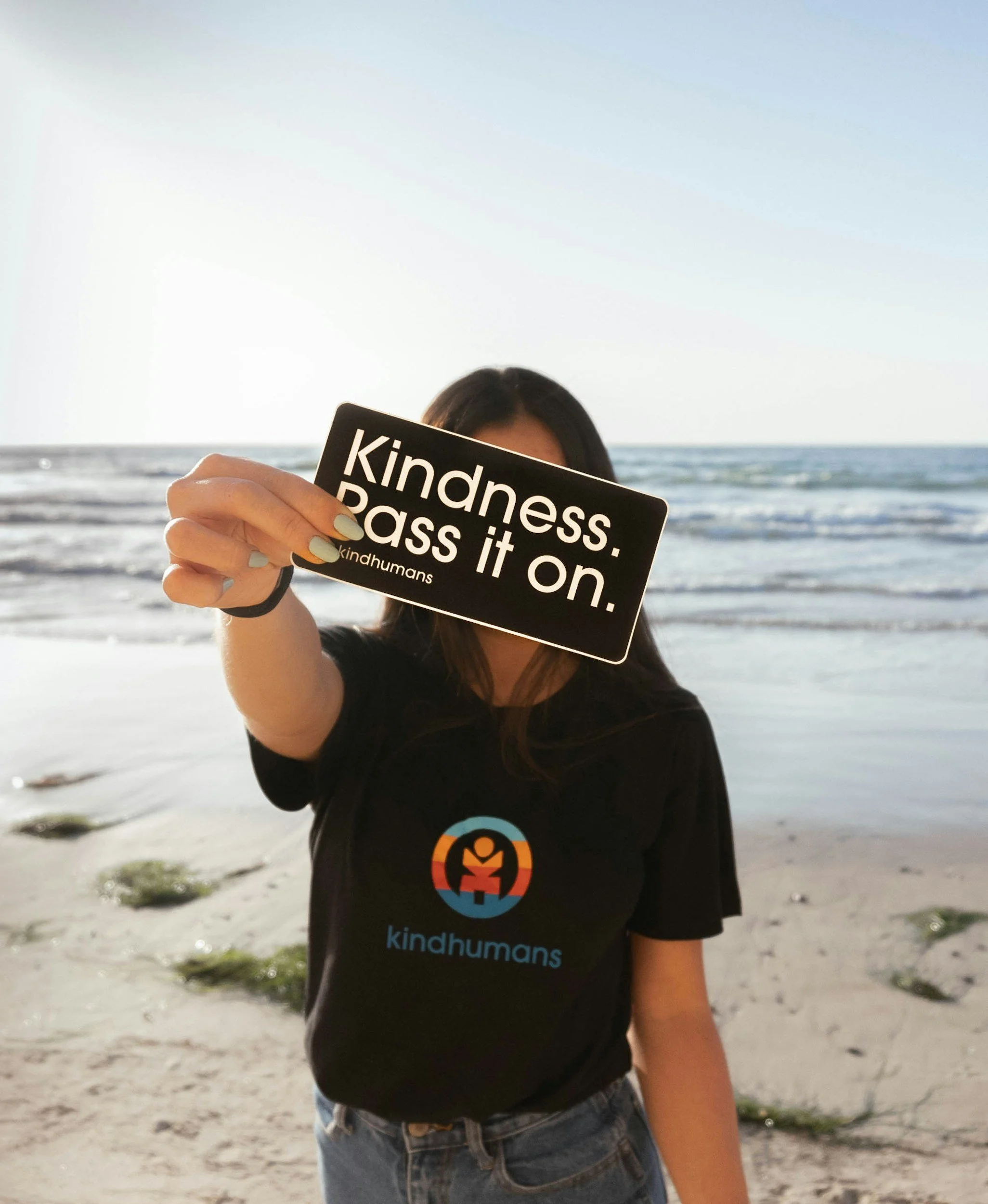 A person holding a black sign that reads 'kindness. pass it on.' in front of their face, standing on a sandy beach with ocean waves in the background. The Data Gap™ impact-led organisations and nonprofit page hero image.