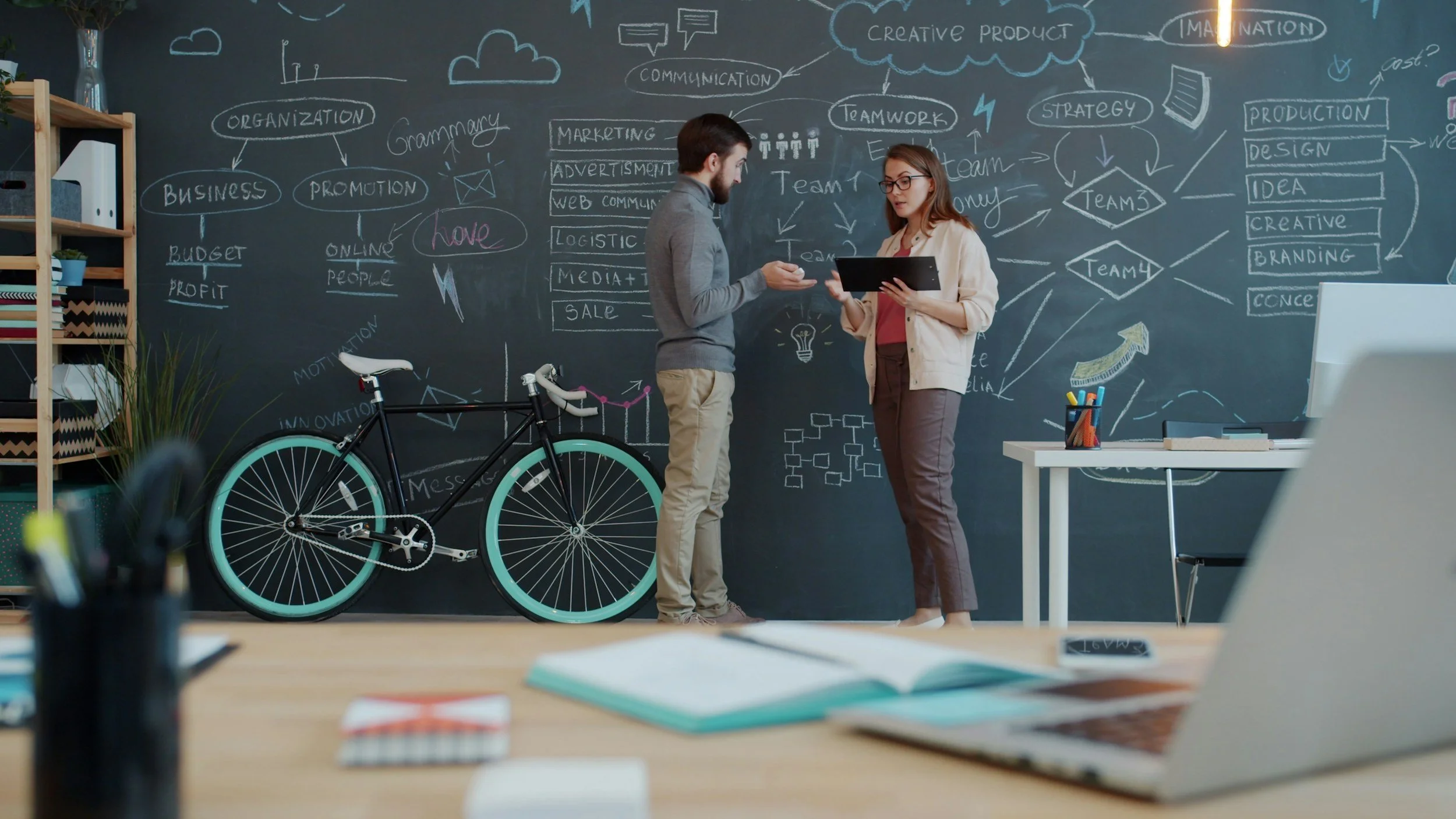 Two people, a man and a woman, standing in front of a large black chalkboard filled with diagrams and notes about business, marketing, and teamwork. There is a bicycle with turquoise wheels leaning against the chalkboard on the left side of the image.