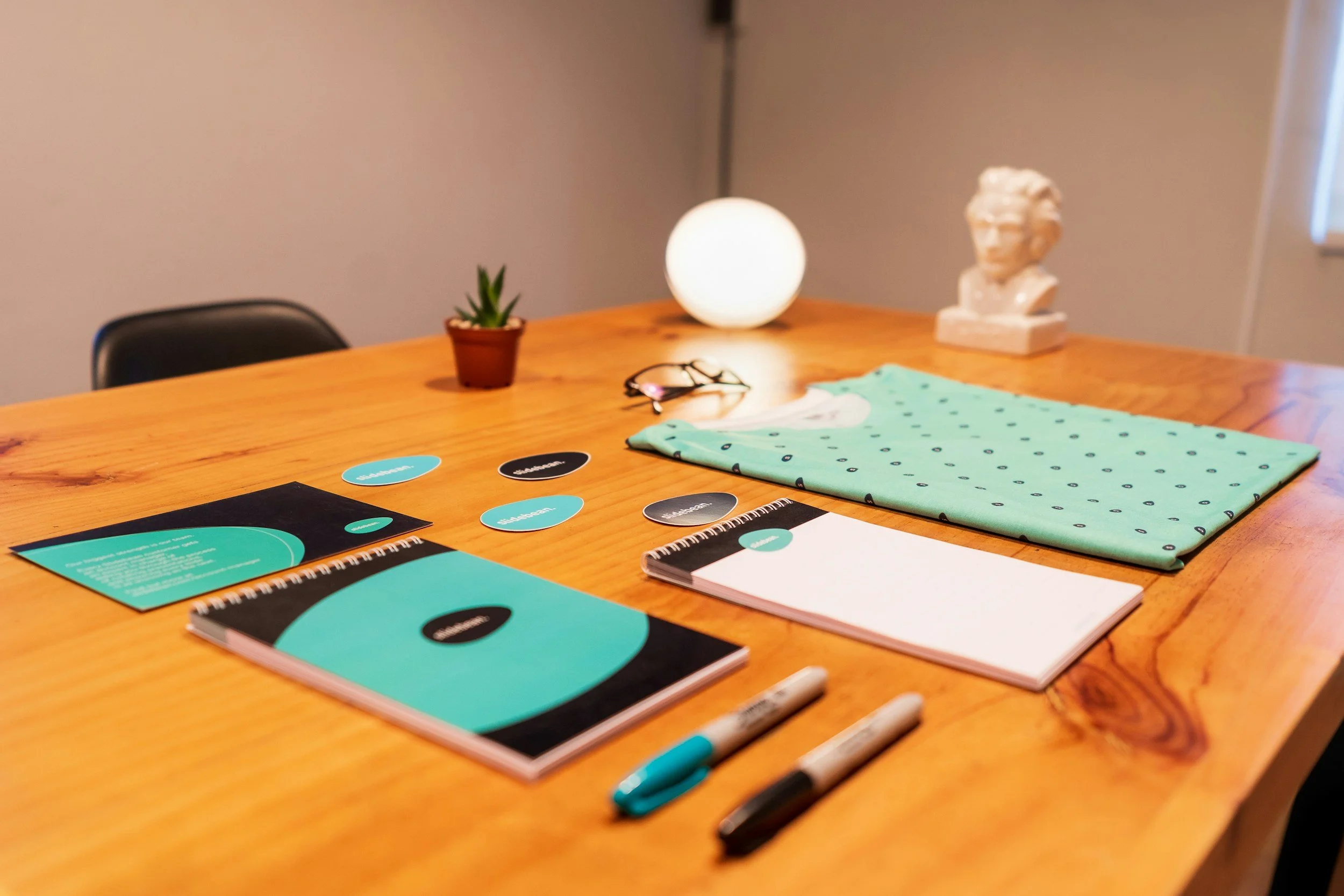 A wooden table holds notebooks, pens, teal and black circular notes with words, a pair of glasses, a folded teal cloth, a small potted plant, a round white lamp, and a bust sculpture in the background.
